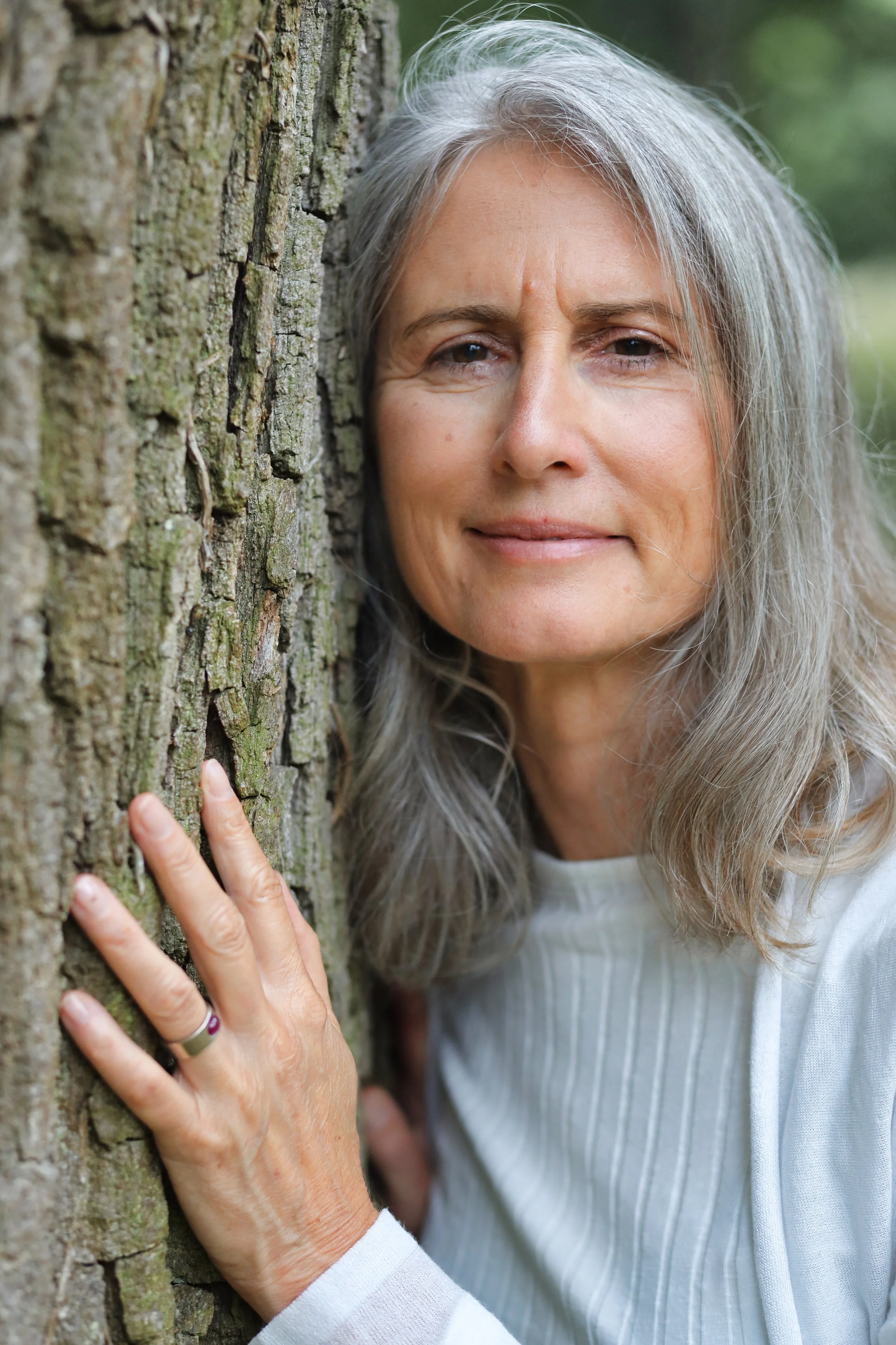 Eine ältere Frau mit grauem Haar lehnt an einem Baum und blickt in die Kamera, umgeben von Natur.