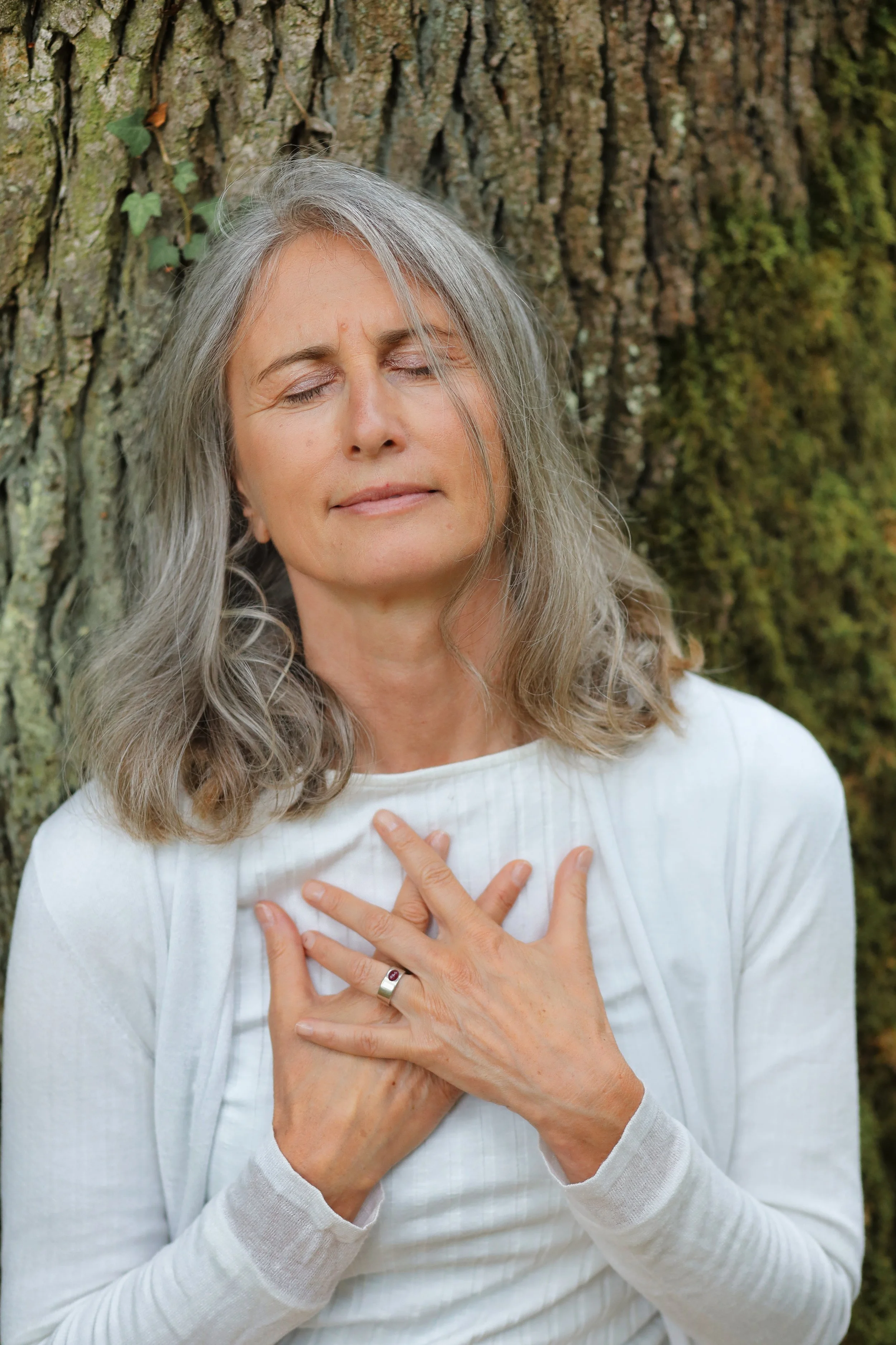 Eine alte Frau mit grauem Haar, die vor einem Baum steht und die Hand auf die Brust legt, mit geschlossenen Augen.