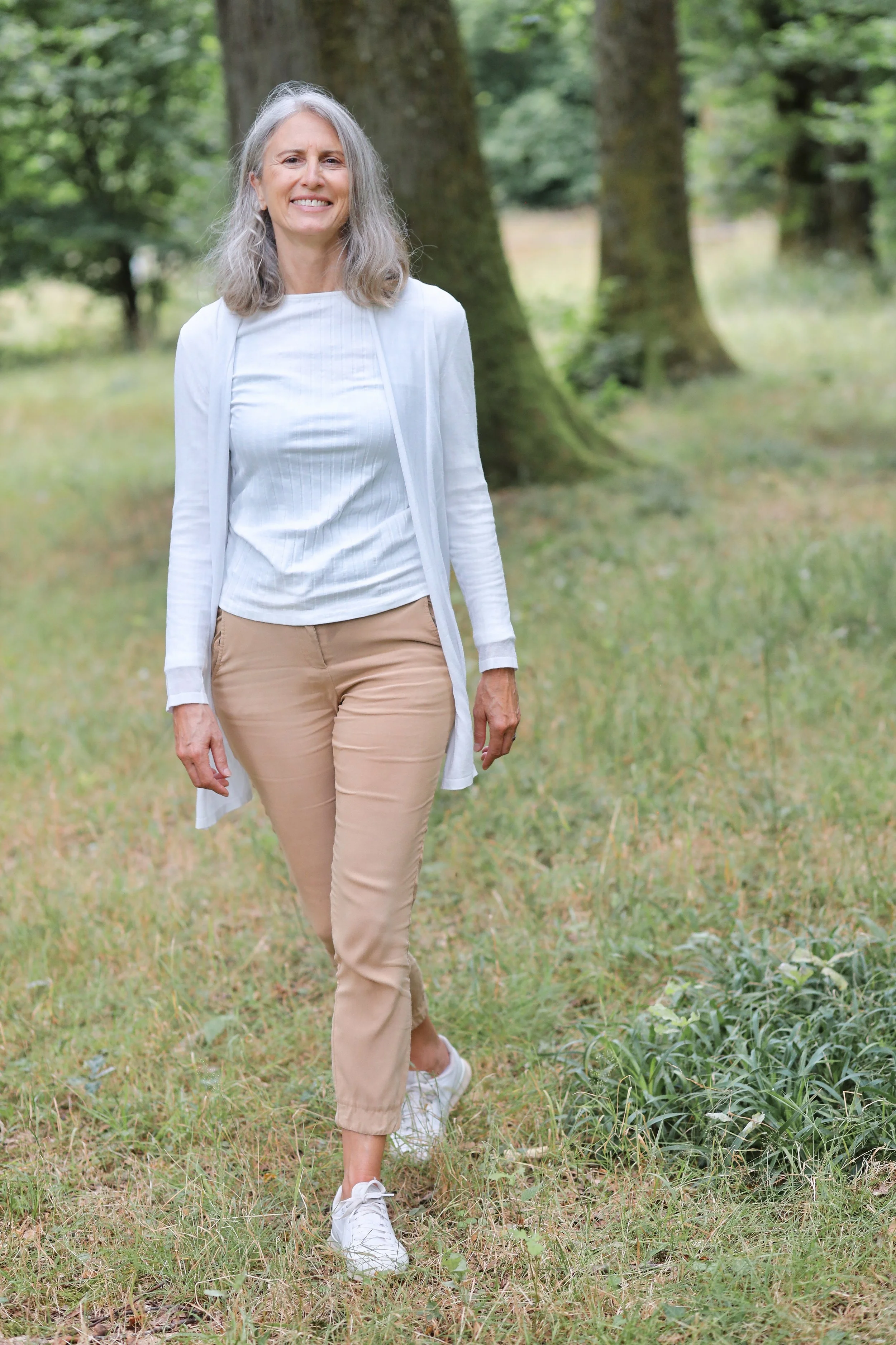 Eine ältere Frau mit grauem, lockigem Haar geht im Park auf einem pfad durch die Natur.