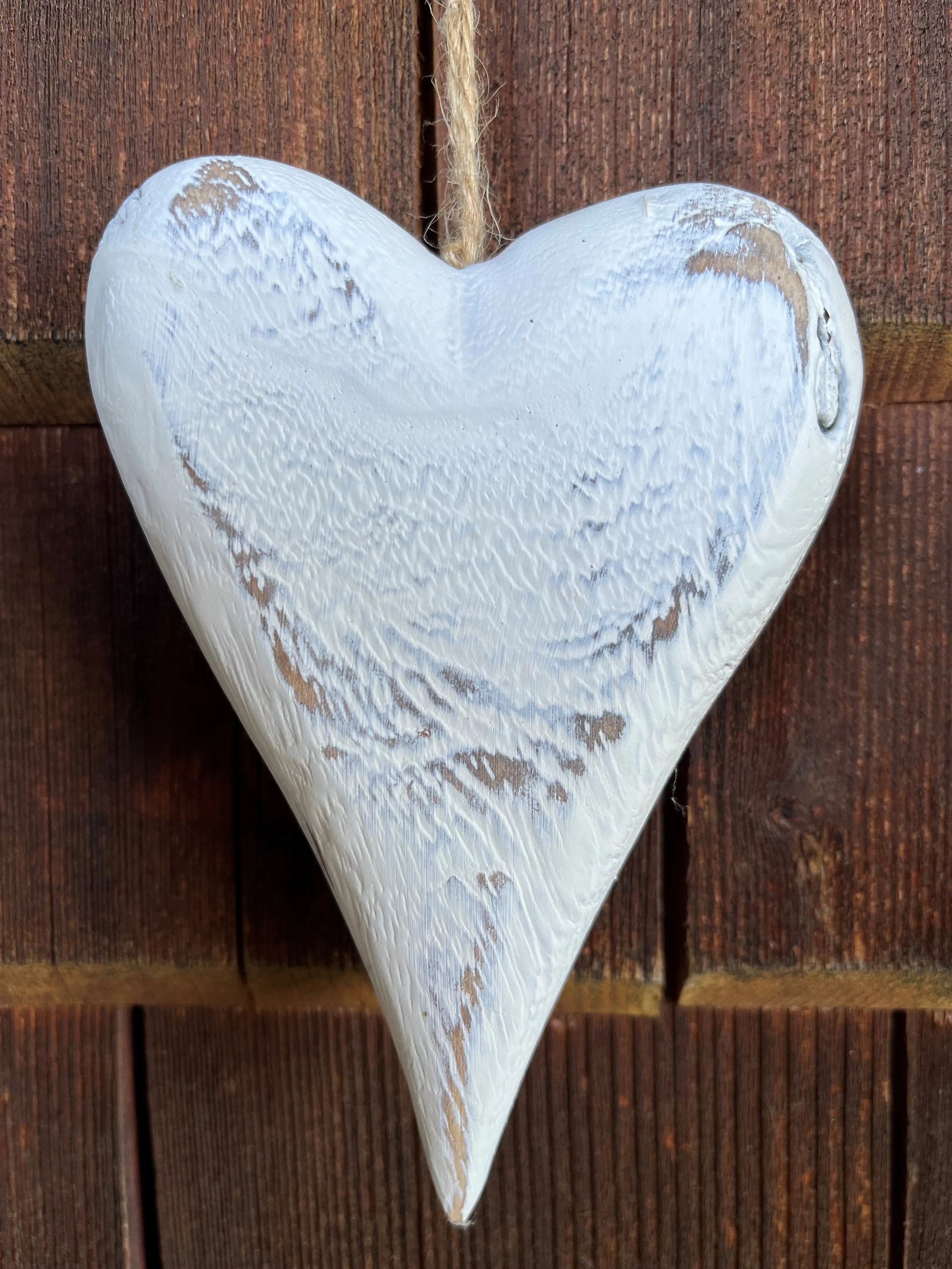 Weißes, herzförmiges Dekorationsstück mit verwittertem Look, hängt an einer Schnur vor einer Holzwand.
