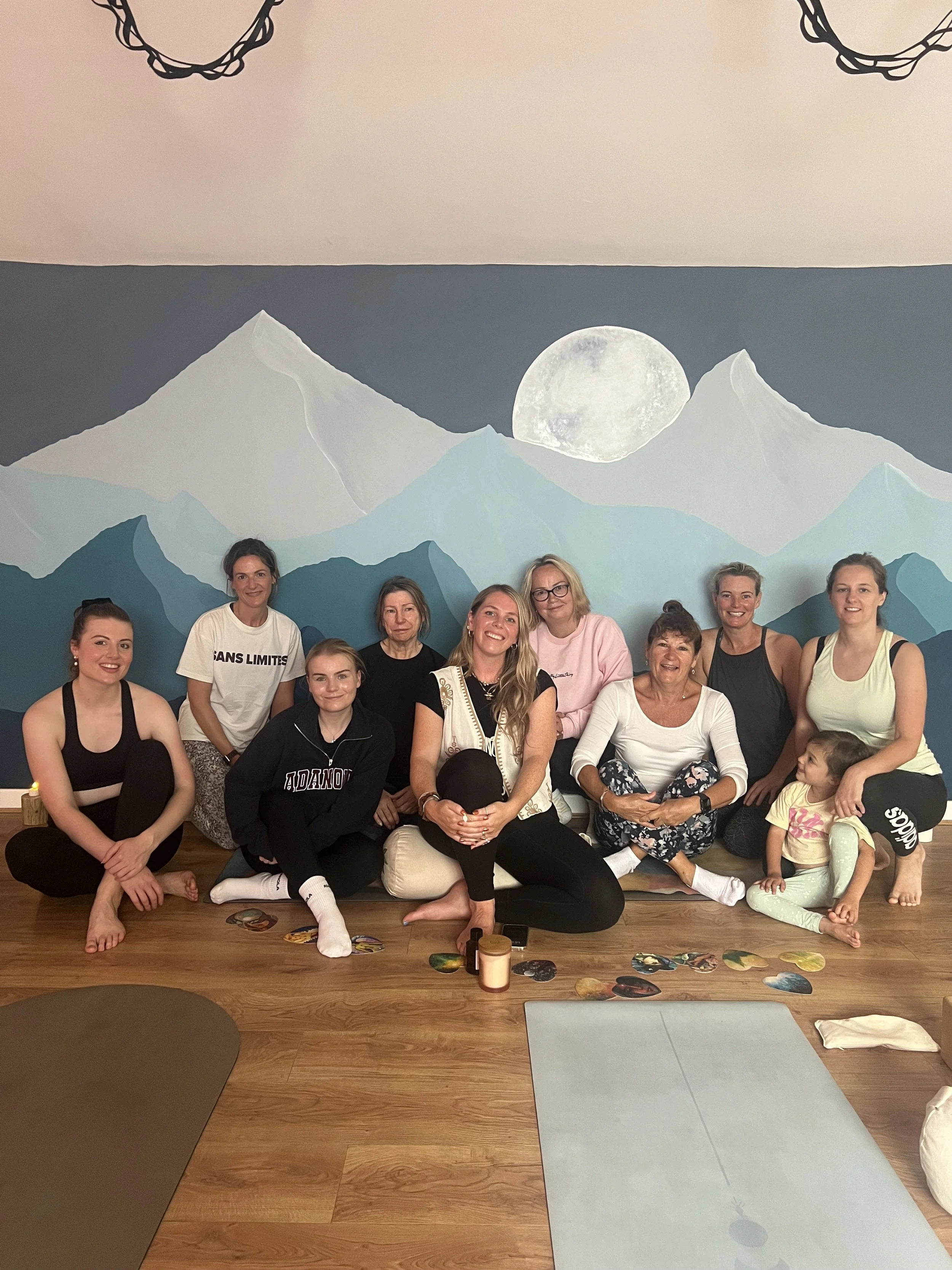 Group of women and a child sitting and kneeling in a yoga or meditation studio after taking part in a community Yoga class with a mountain and moon mural background, yoga mats, and scattered decorative objects.