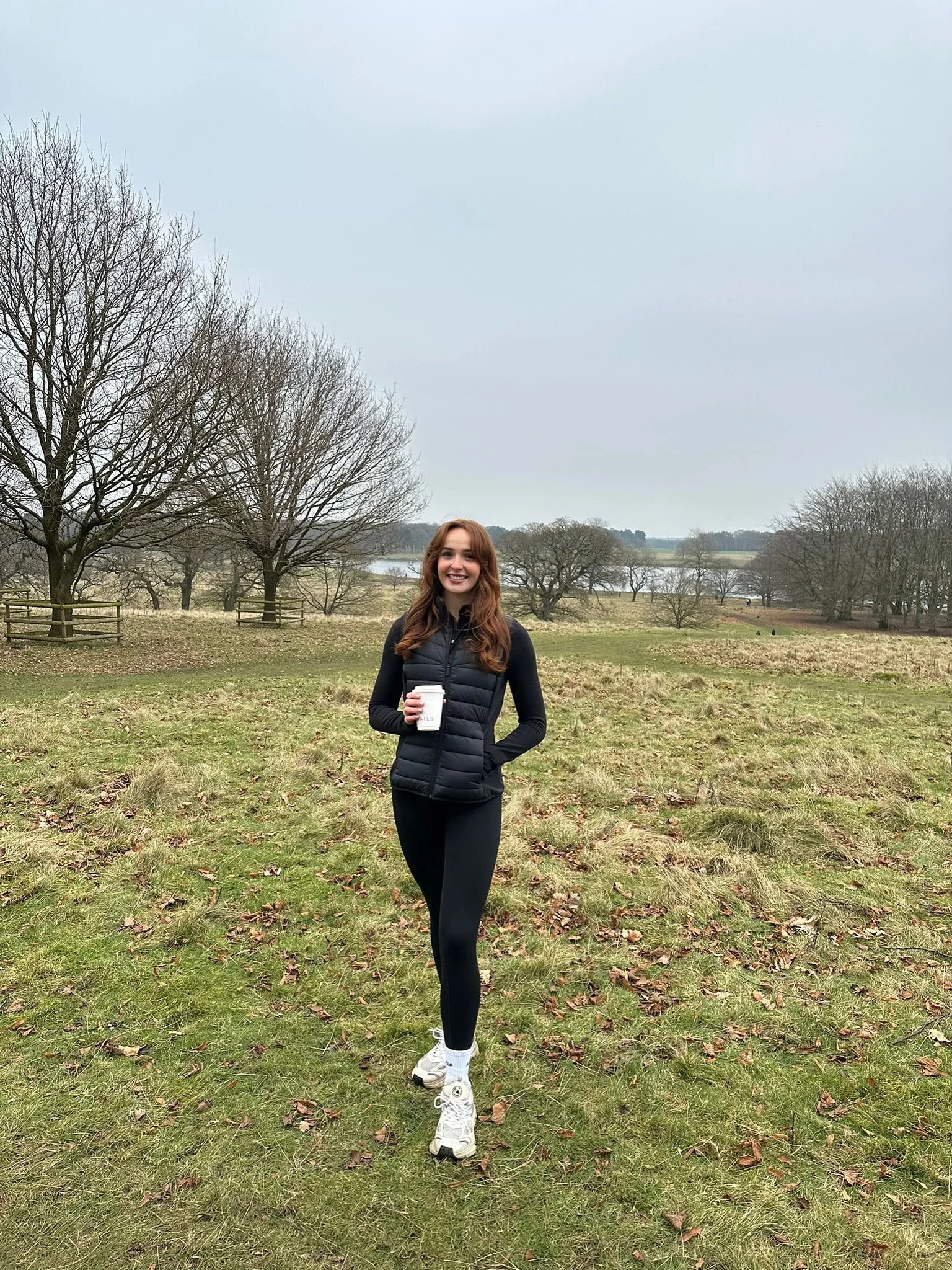 A young woman with long brown hair smiling, holding a white coffee cup, standing outdoors in a grassy field with trees and a lake in the background on a cloudy day.