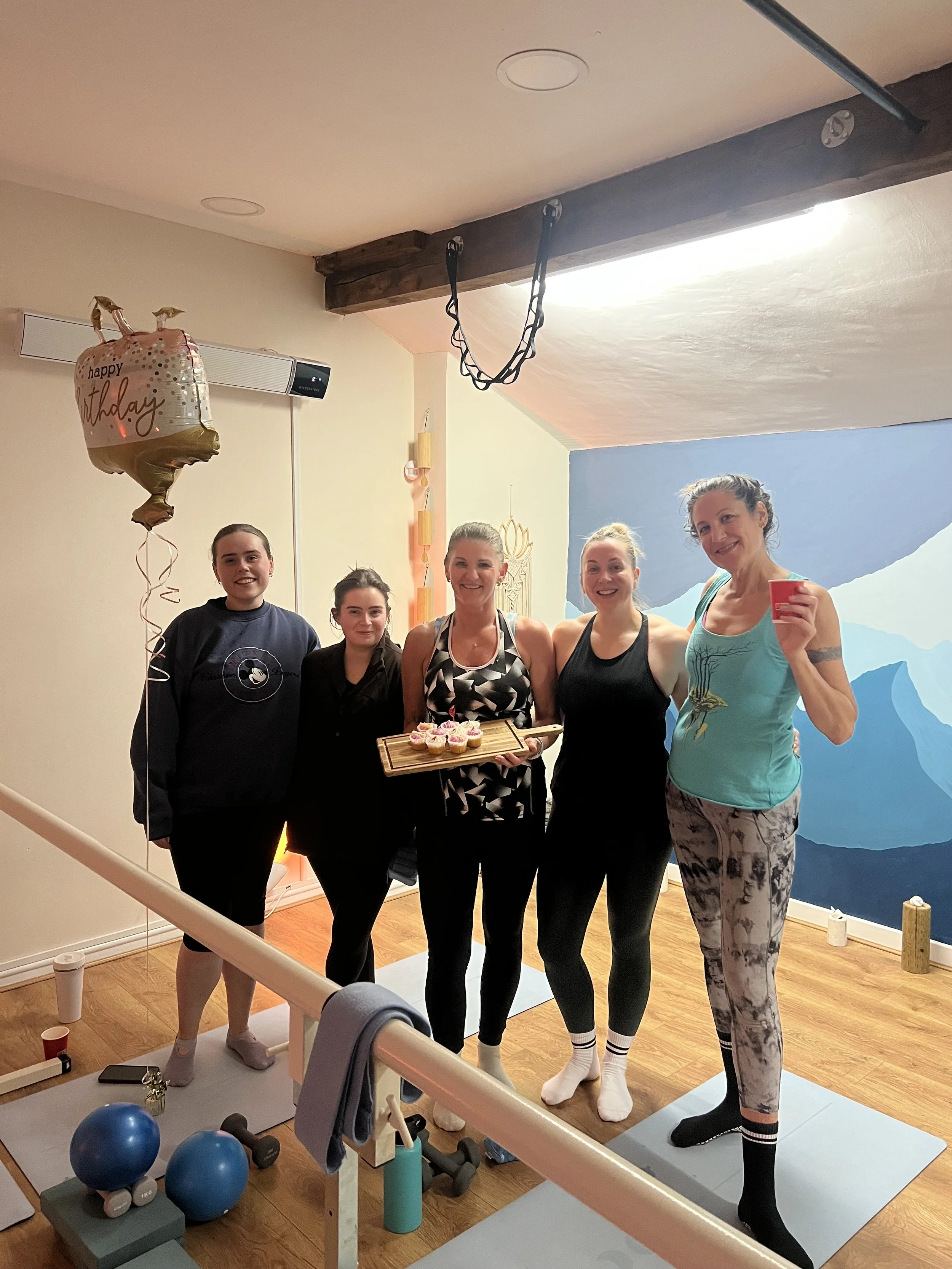 Group of five women in workout clothes celebrating a birthday with cupcakes inside a fitness studio, with a heart-shaped balloon saying 'happy birthday' visible in the background.