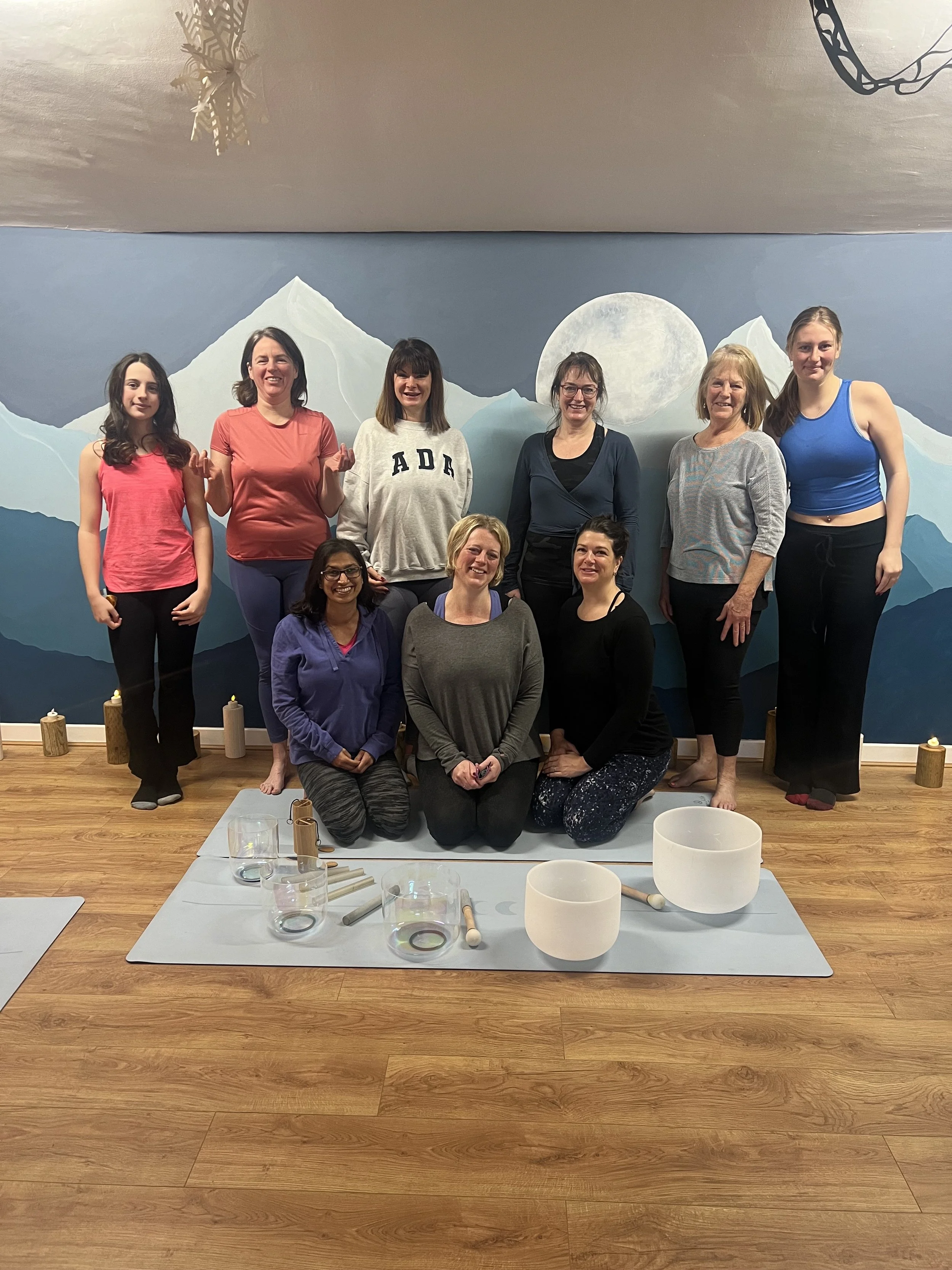 A group of nine women in a yoga studio, some sitting and others standing, with a mountain and moon mural in the background, and yoga mats, singing bowls, and candles on the floor.