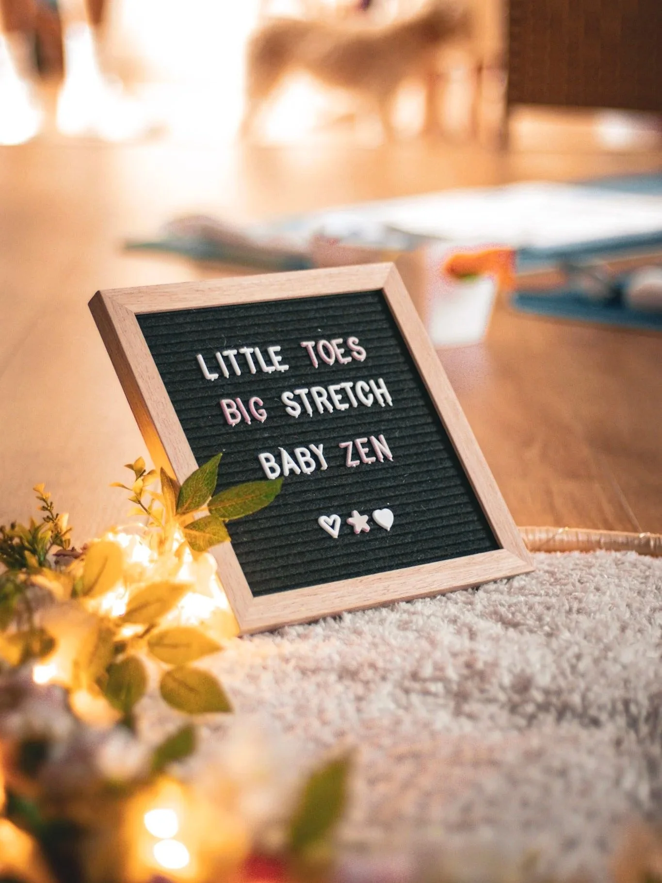 A framed letter board on a table with a decorative sign that says 'Little Toes Big Stretch Baby Zen,' surrounded by small flowers and warm lighting.