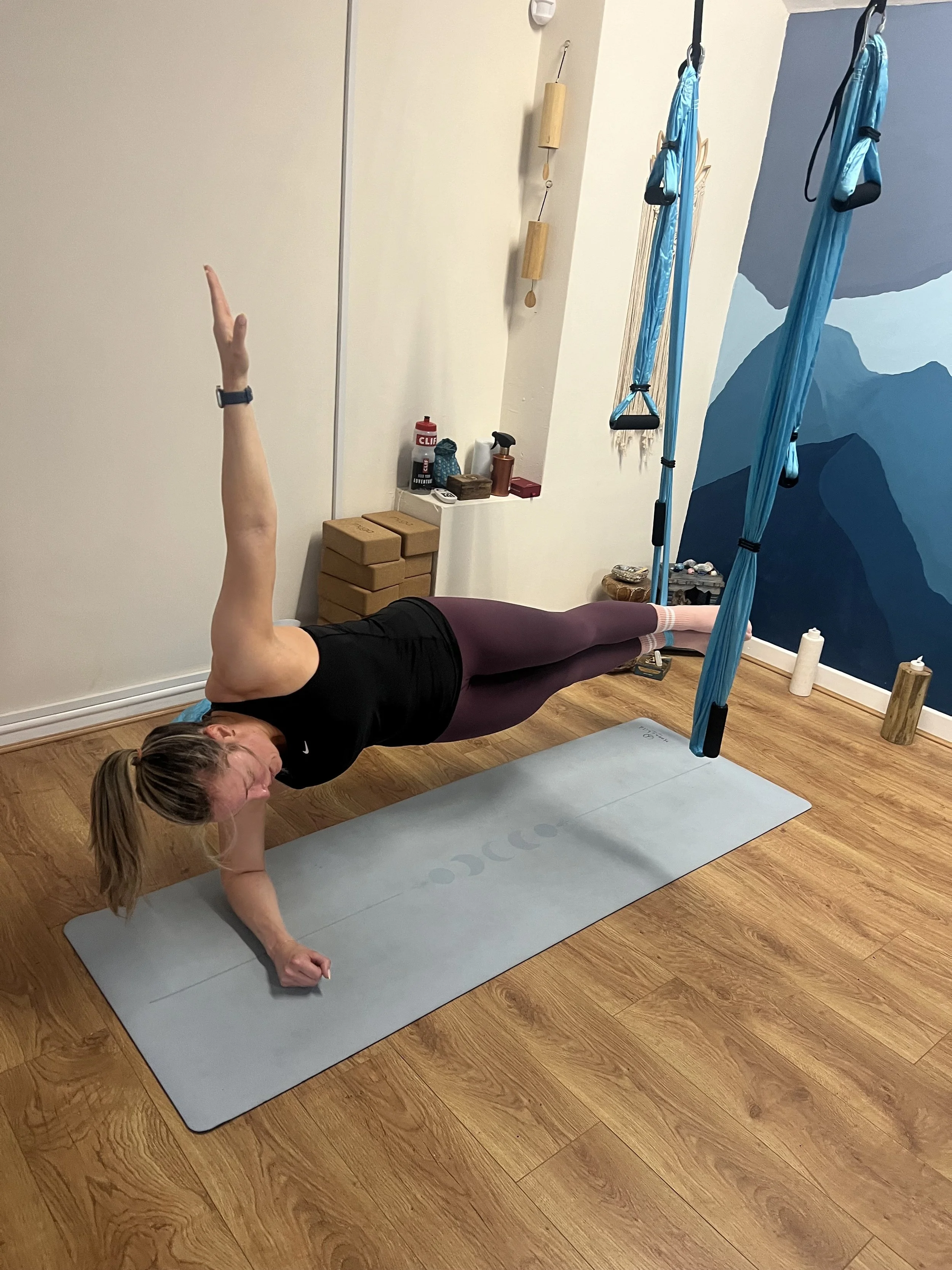 A woman performing a one-arm balance yoga pose on a yoga mat in a room with wooden floors. She is supporting herself on her left arm with her body parallel to the floor, her right arm raised, and her legs extended behind her. Exercise equipment and blue yoga hammocks hang from the ceiling.