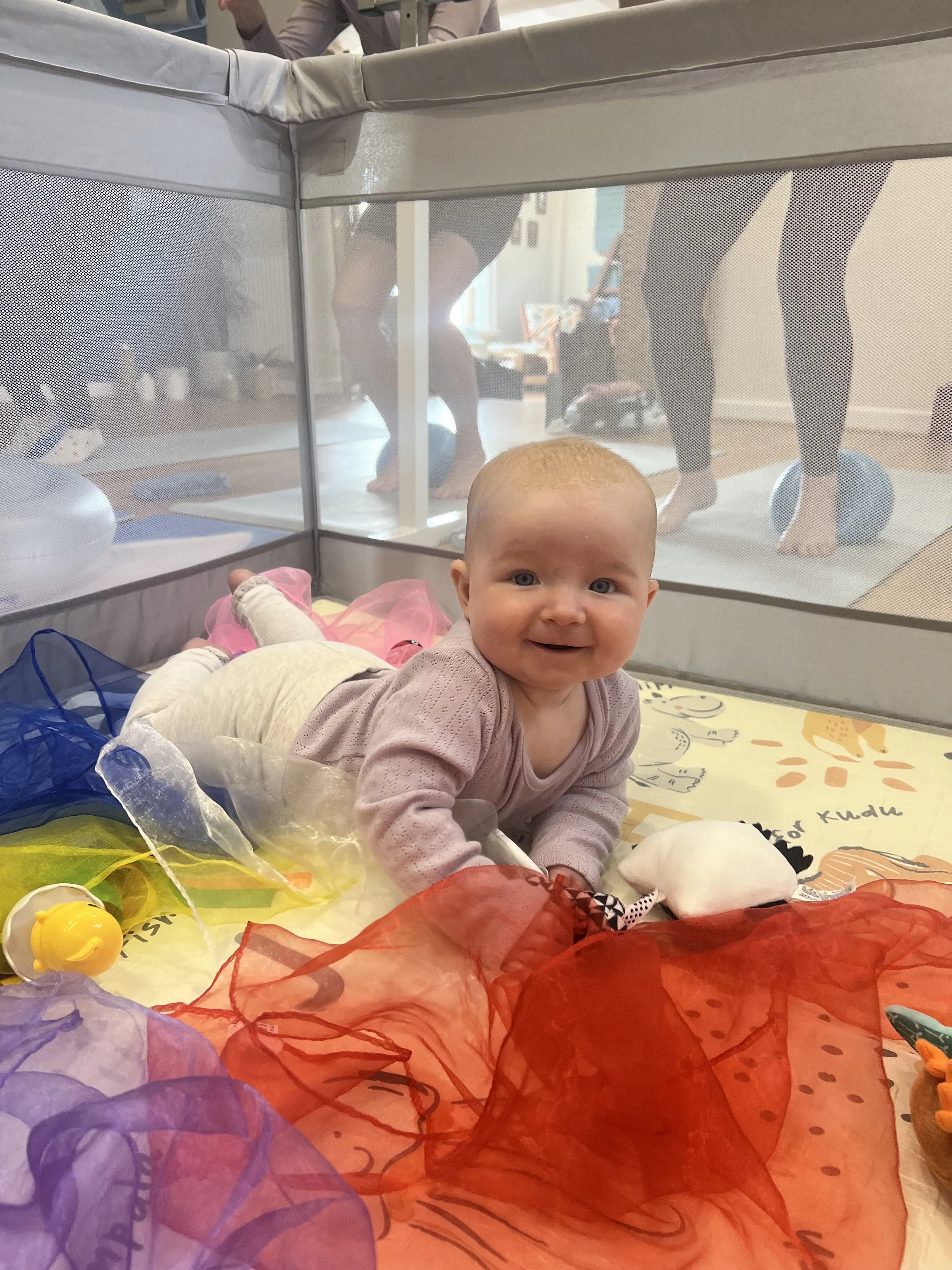 A baby crawling on a playmat inside a playpen, smiling at the camera. The playmat has colorful fabrics and toys. In the background, people are taking part in a Mamma Barre class, a Barre class where you can bring your baby with you. 