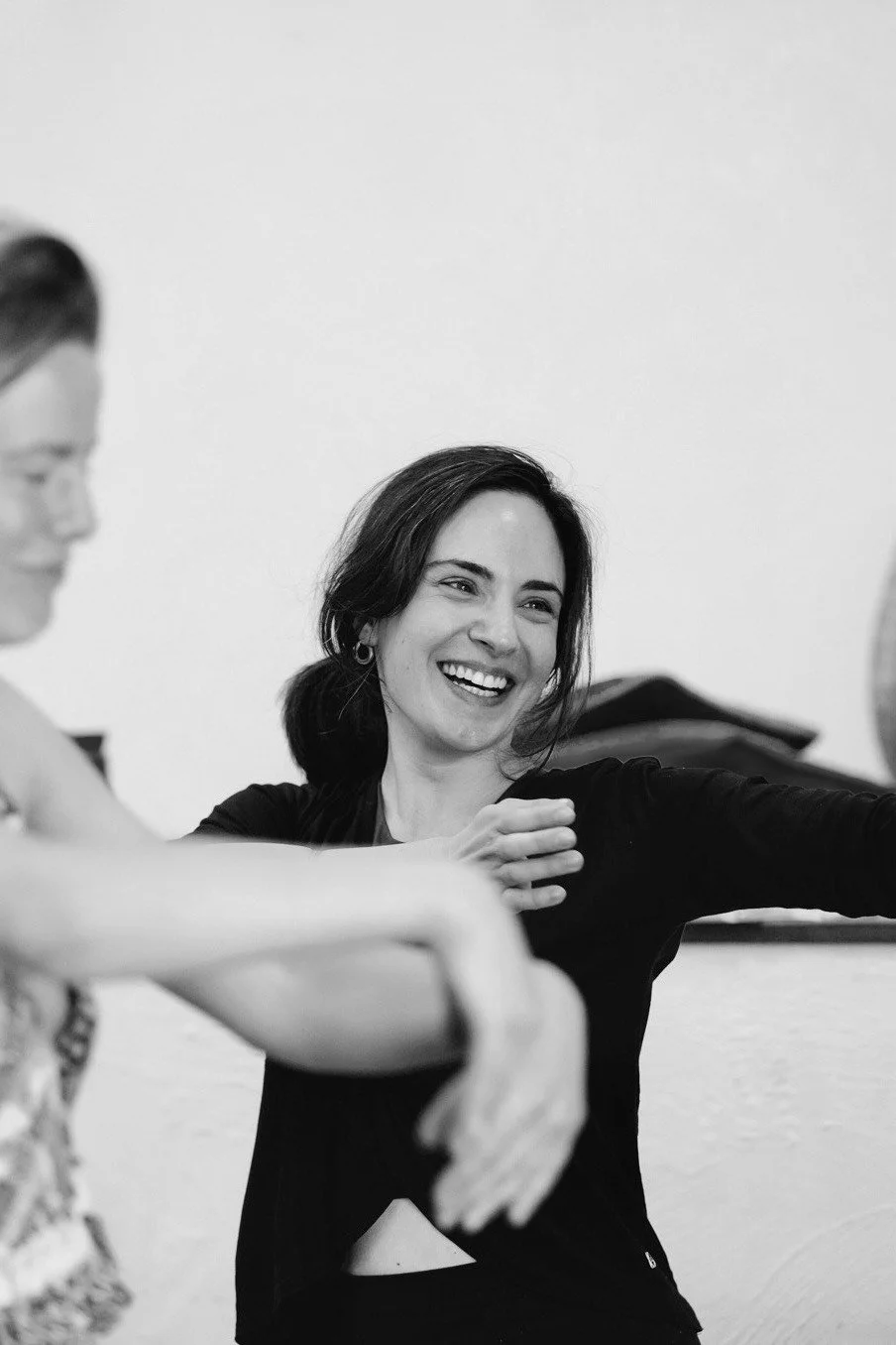 A woman smiling and stretching during a fitness or dance class, with another person partially visible nearby.