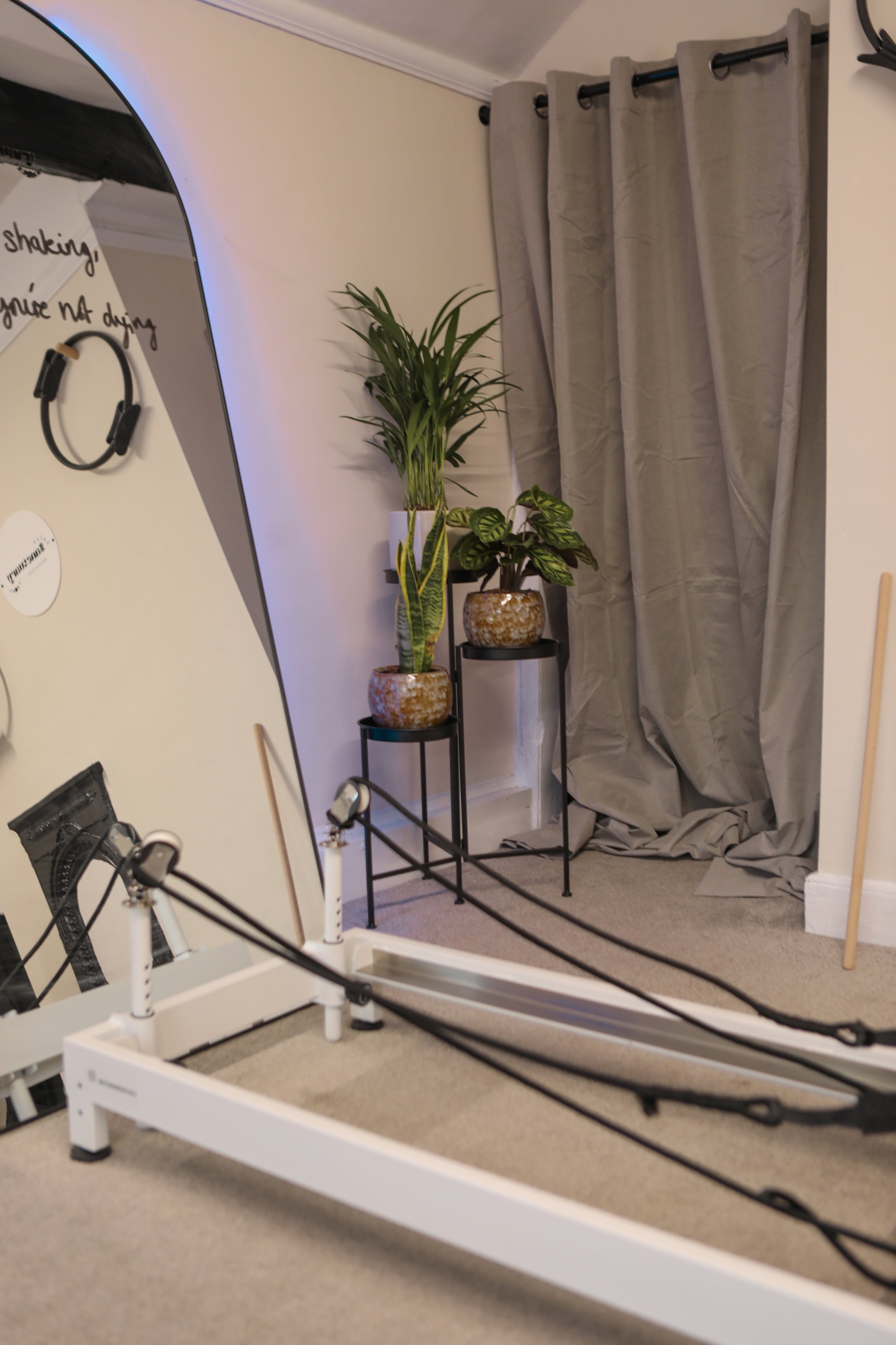 A Pilates studio with two reformer machines, a large mirror leaning against the wall, a small black cabinet, and wall art. The room has beige walls, a window, and decorative blue lighting.