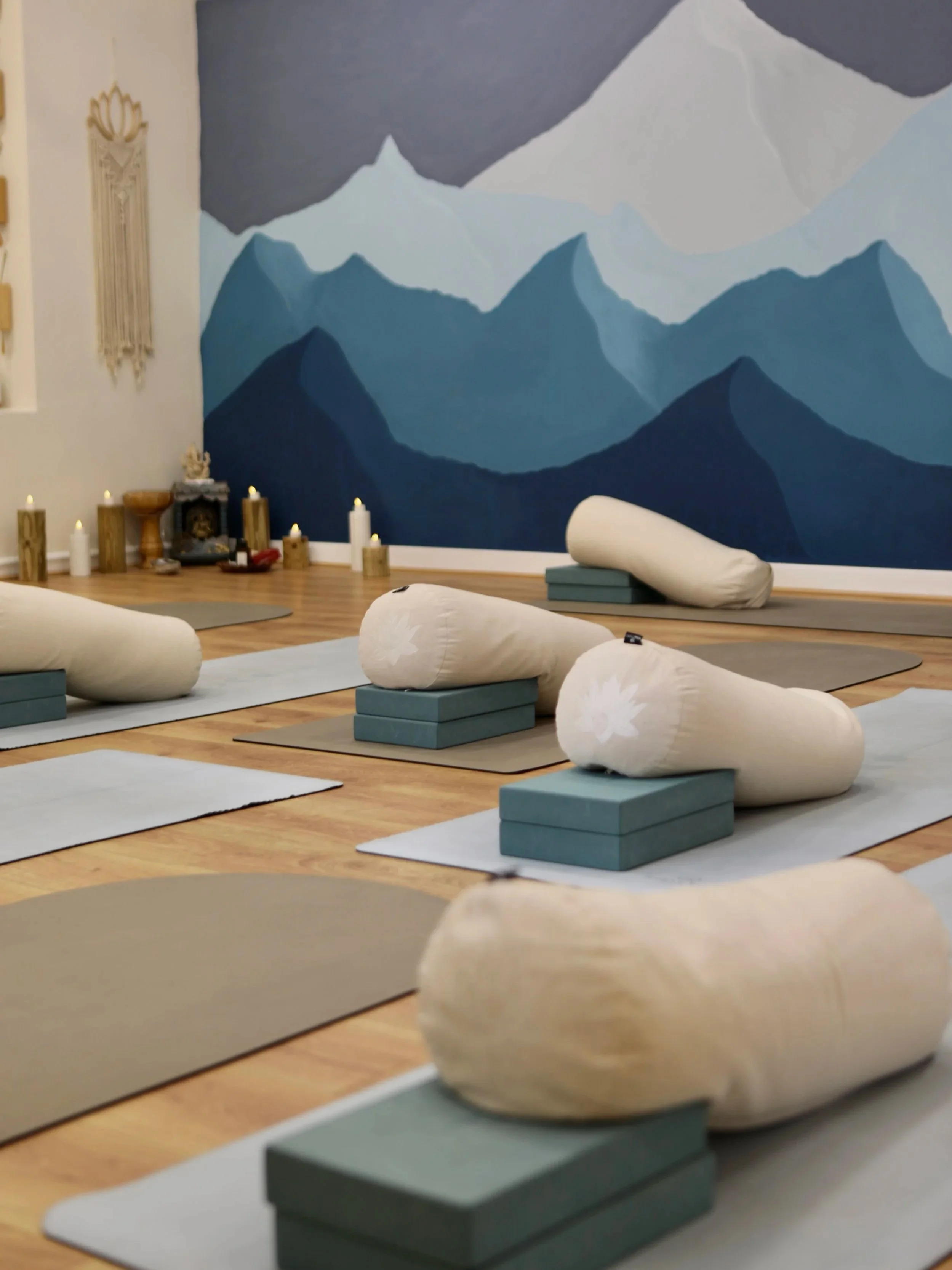 A yoga studio with folded bolsters and mats arranged on a wooden floor, potted candles, and a mountain mural on the wall.