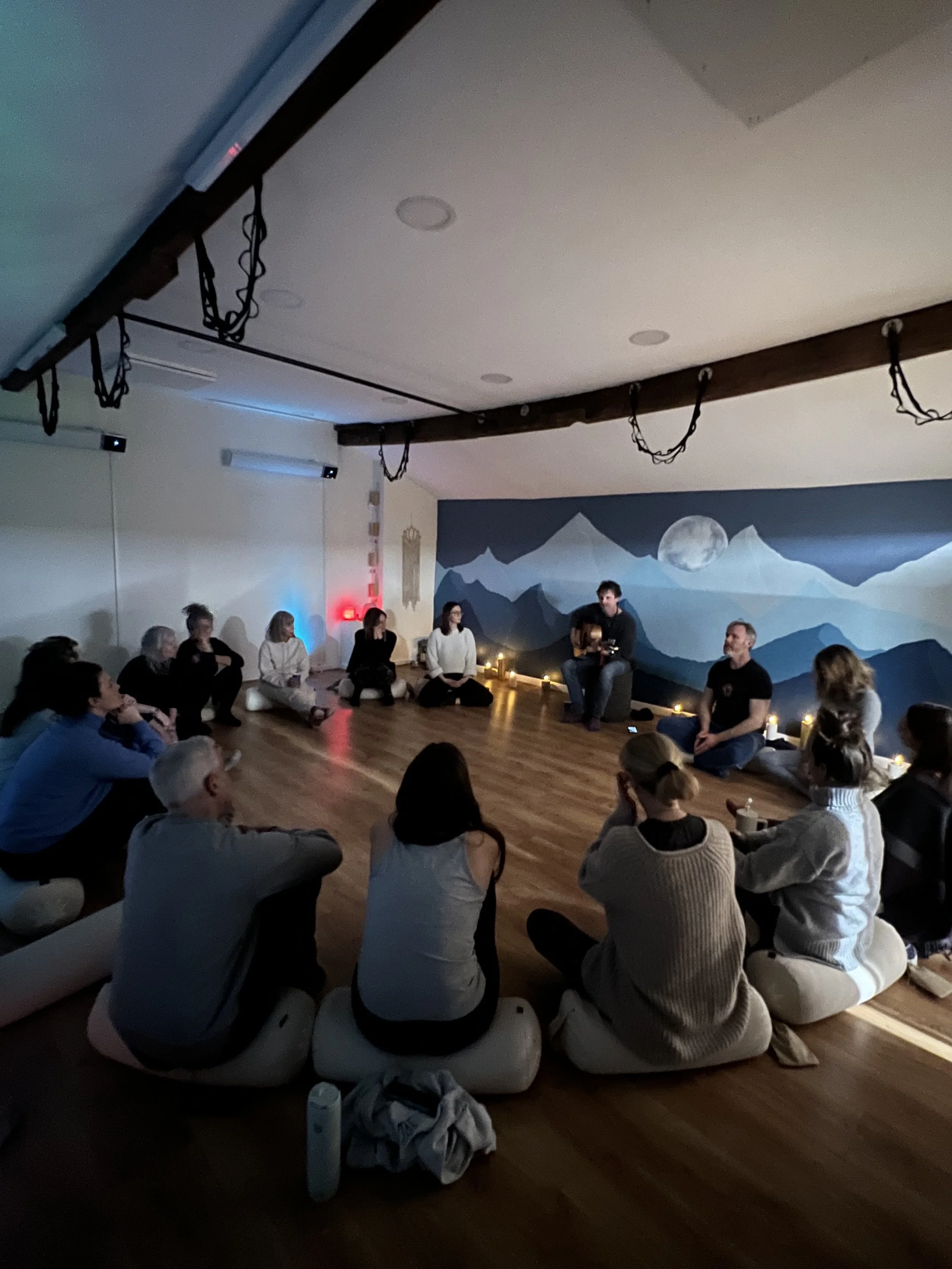 People sitting in a circle in a cozy room with a mural of mountains and a full moon on the wall, participating in a group Kirtan. Candles and soft lighting create a calming atmosphere.