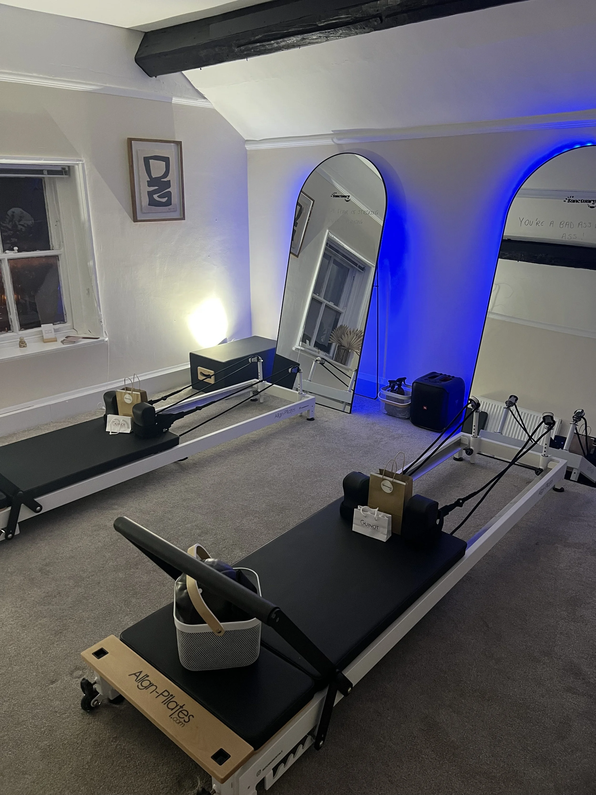 A Pilates studio with two reformer machines, a large mirror leaning against the wall, a small black cabinet, and wall art. The room has beige walls, a window, and decorative blue lighting.