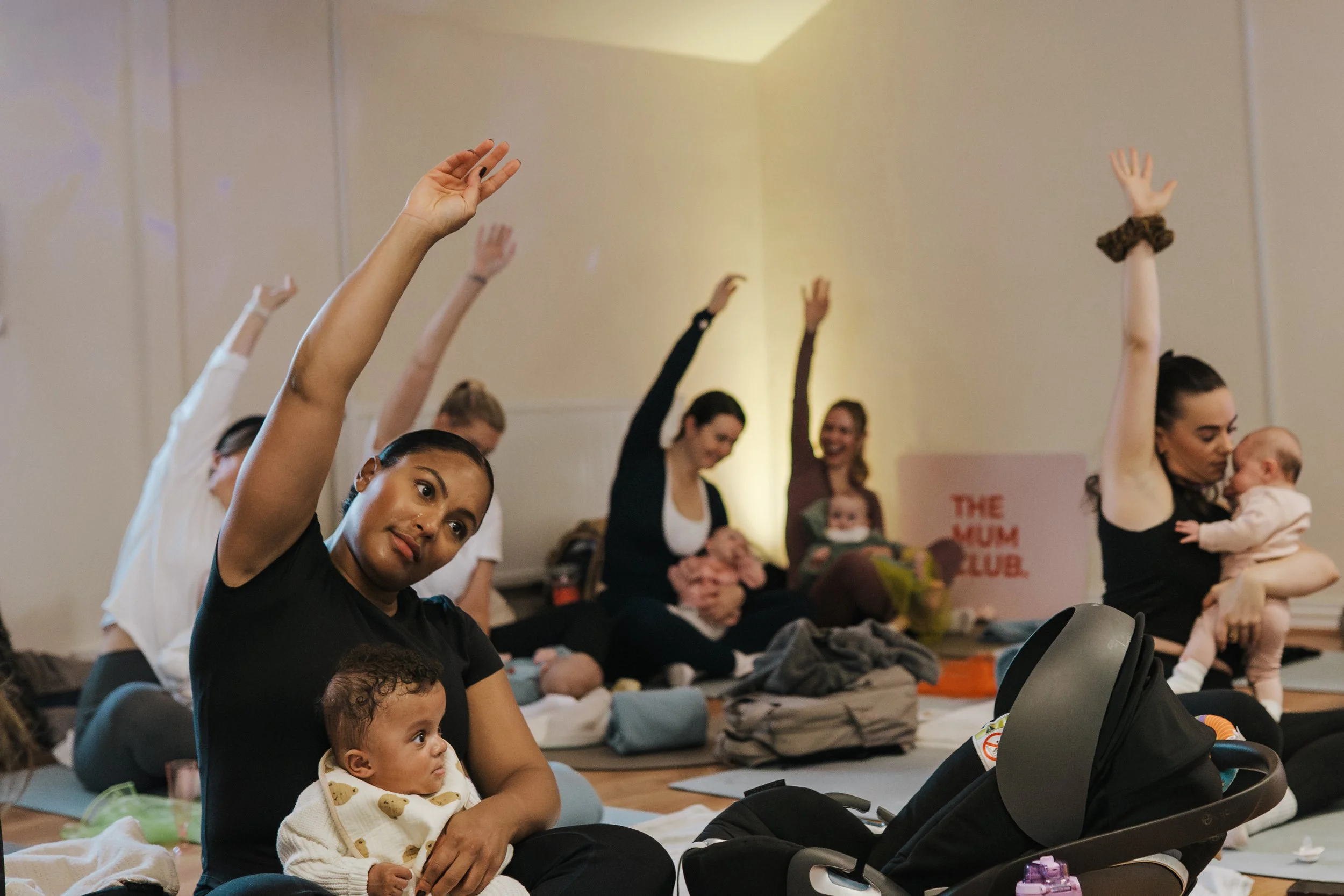 Women and babies participating in a prenatal or mother and baby yoga class, seated on mats with legs crossed, raising their hands. Some women are holding their babies, and there is a baby stroller in the foreground. The atmosphere appears relaxed and