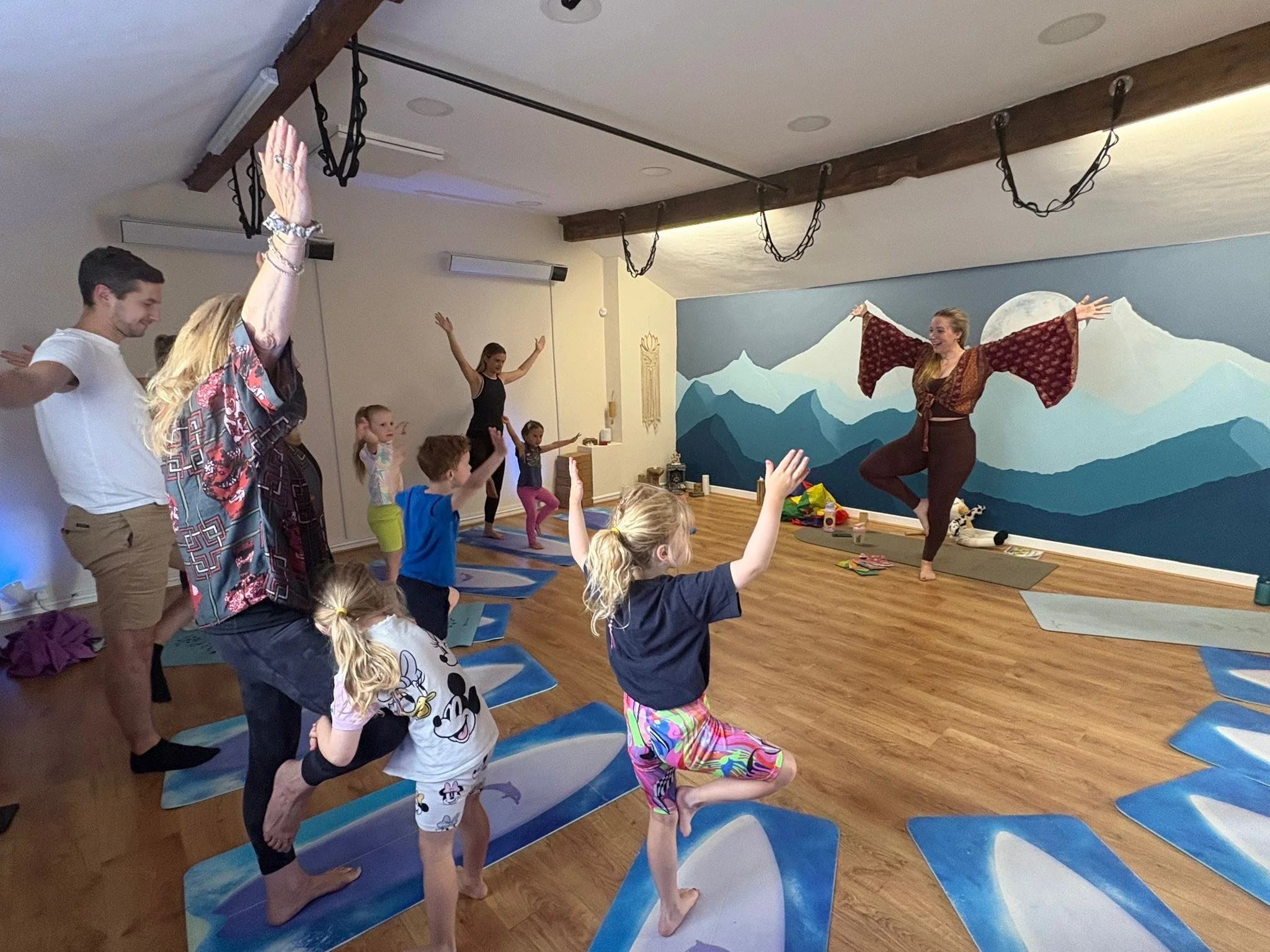 Children and adults participating in a yoga class in a studio with a mountain mural wall, practicing poses with an instructor leading at the front.