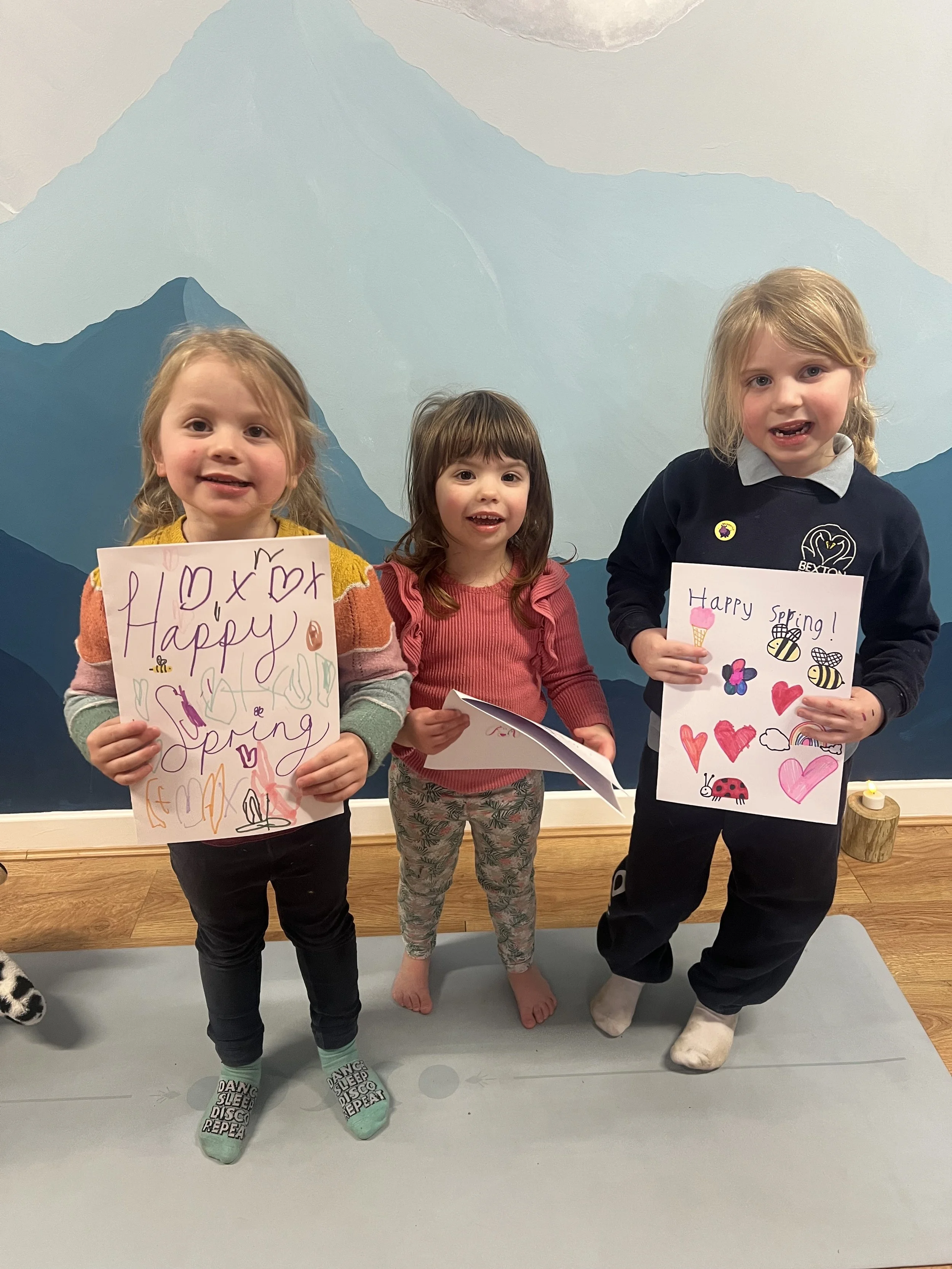 Three young girls standing indoors in front of a mountain mural, holding handmade Valentine's Day or spring greeting cards decorated with hearts, bees, butterflies, and rainbows.