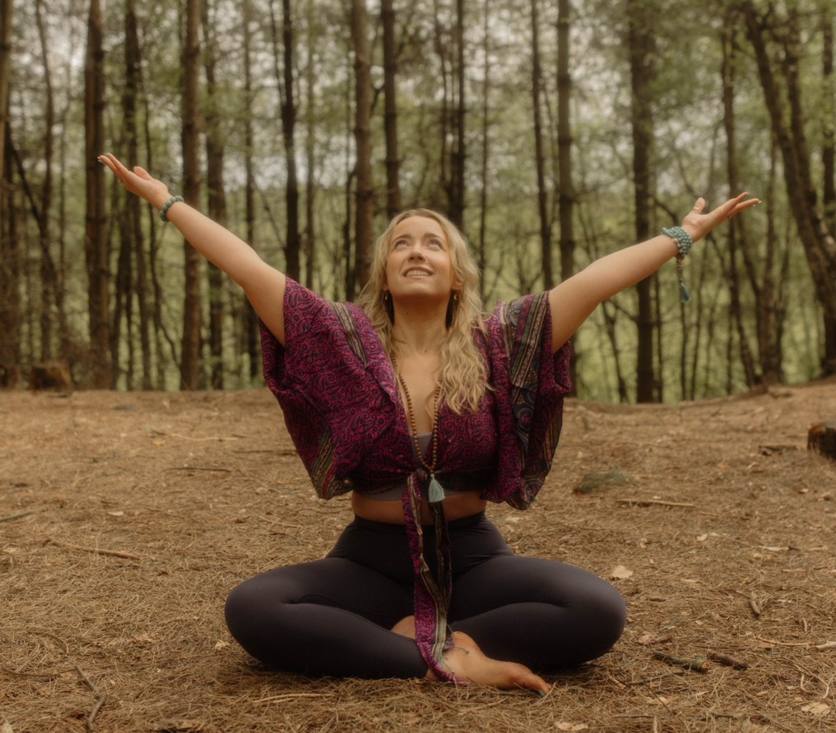 A woman practicing yoga outdoors in a forest with arms extended upward, sitting cross-legged on the ground.