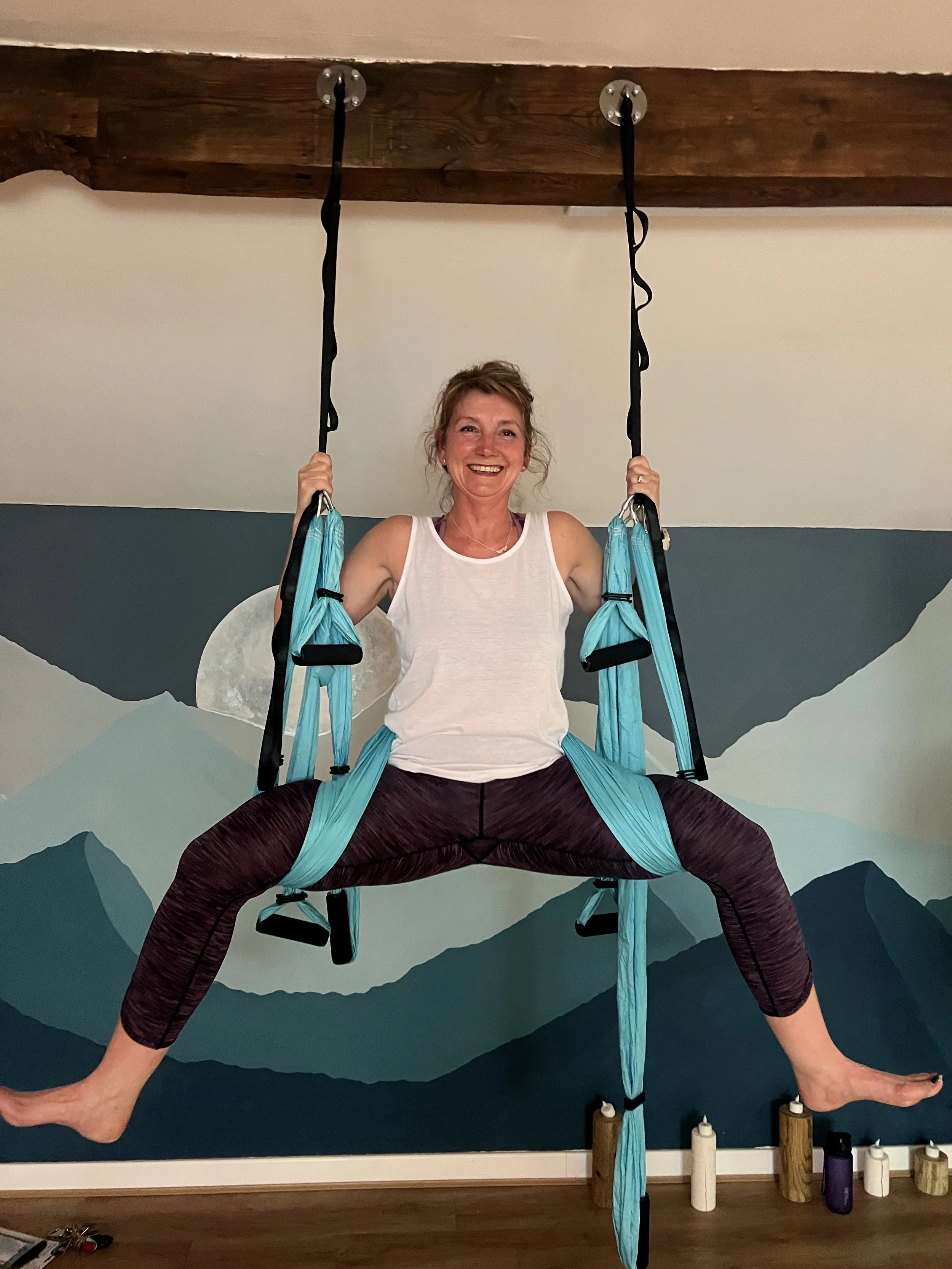 A woman practicing Trapeze yoga using a hammock suspension system in a room with mountain mural wall art.