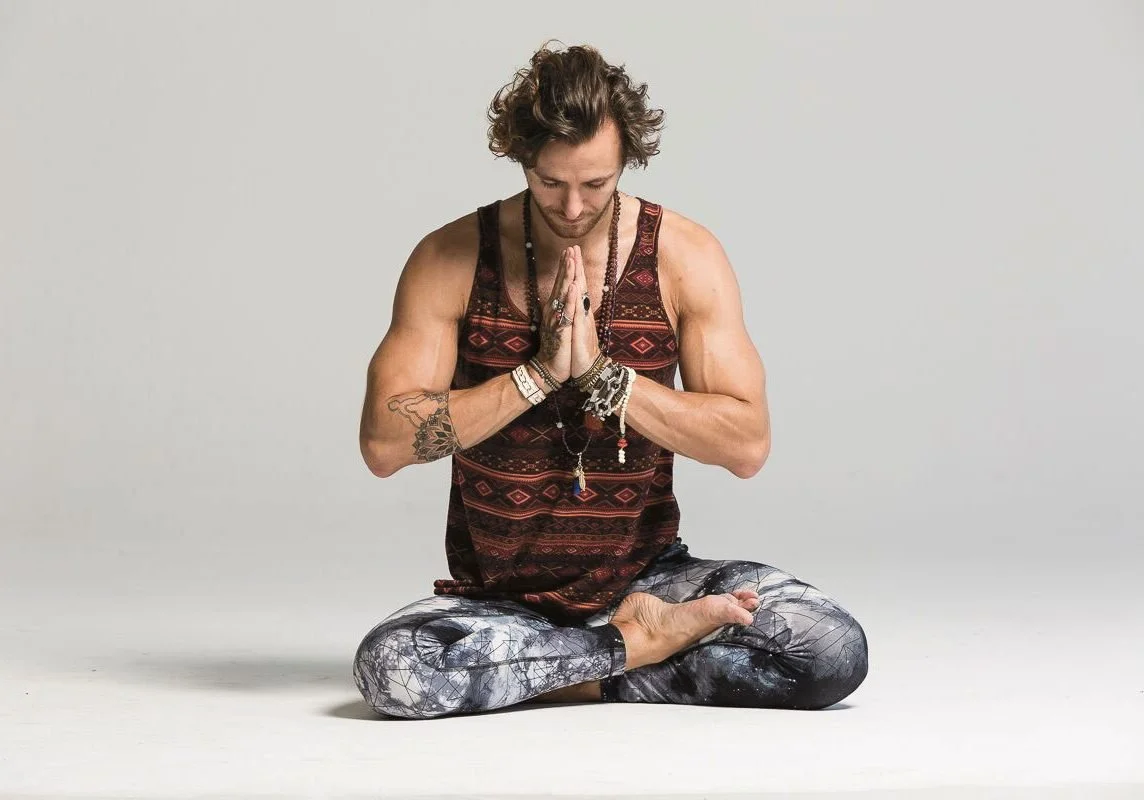 A man practicing yoga in a seated, cross-legged position with hands pressed together in prayer pose, wearing a sleeveless patterned tank top and leggings with a galaxy print, against a plain light background.