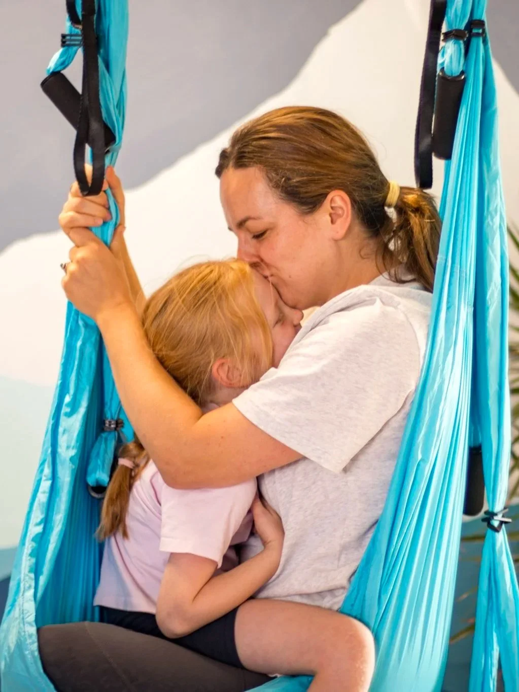 A woman and young girl share a tender kiss while sitting in a blue Trapeze Yoga hammock, capturing a moment of affection.