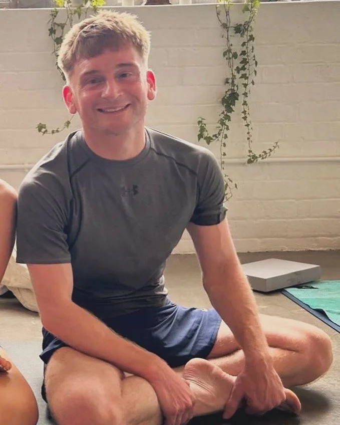 A young man with short blond hair sitting cross-legged on the floor, smiling at the camera, wearing a gray athletic shirt and navy shorts, with a background of a white brick wall and some green climbing plants.
