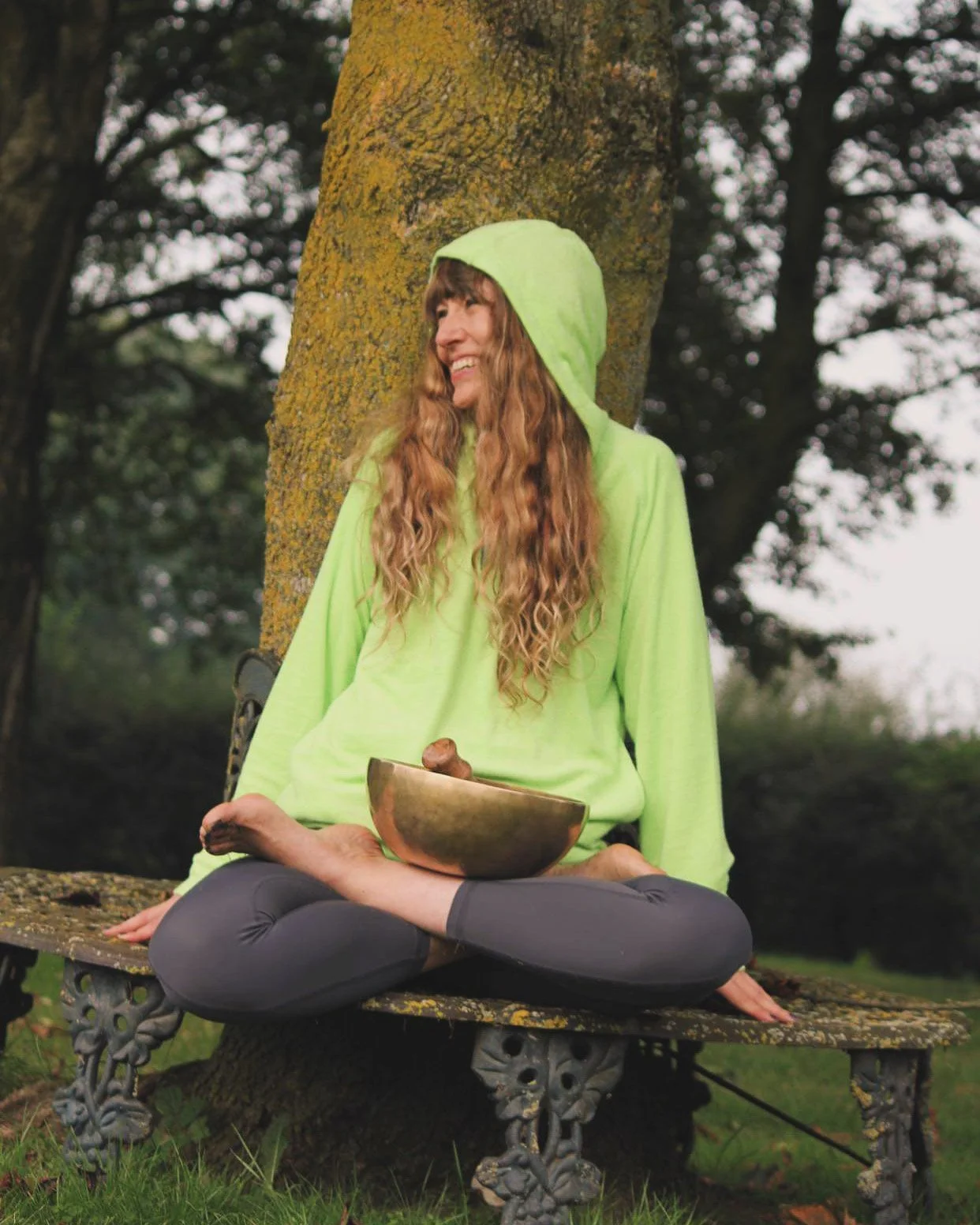 A woman with long wavy hair wearing a bright green hoodie, sitting cross-legged on a park bench, holding a bowl and smiling, with trees in the background.