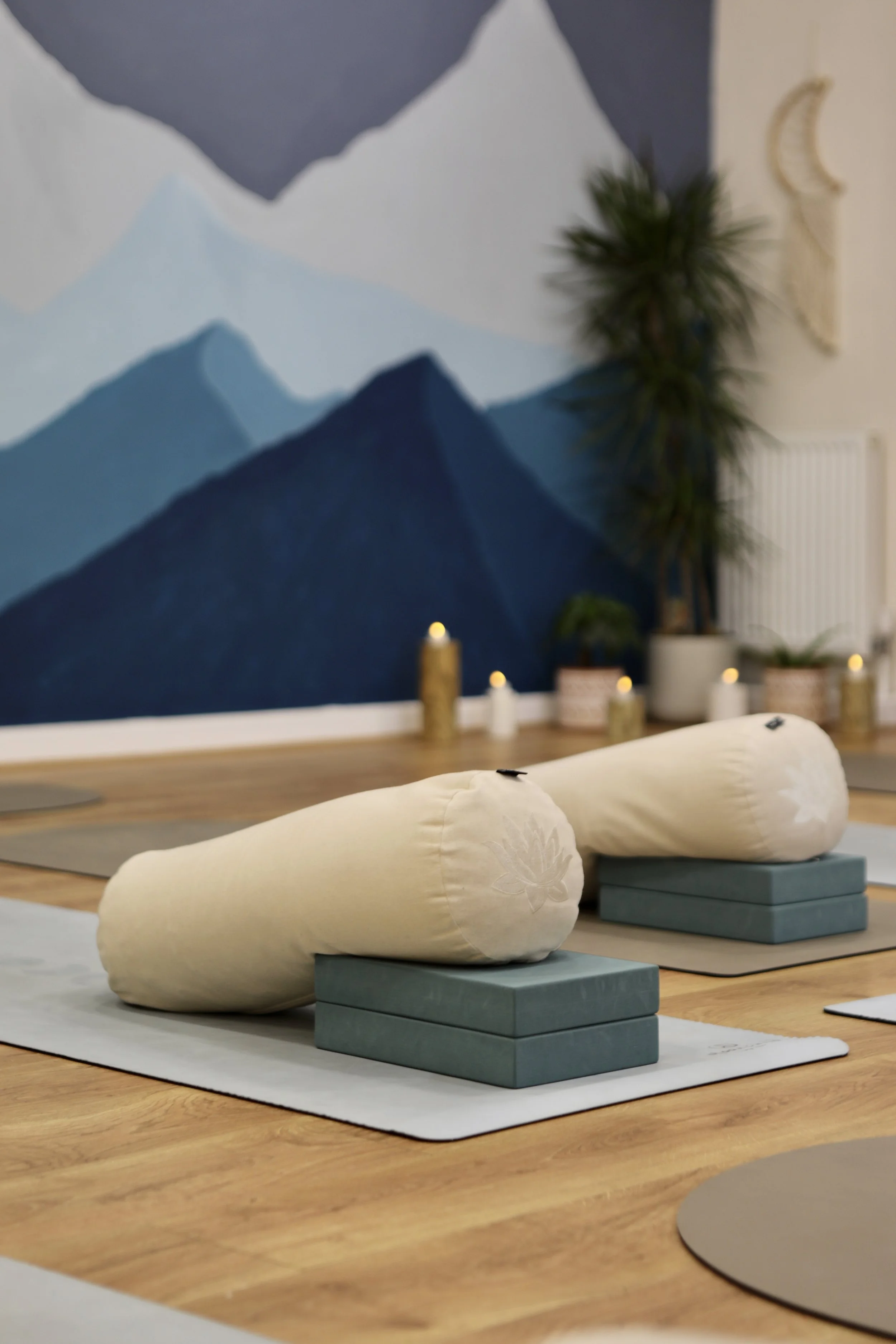 Yoga props including bolsters and blocks set on mats in a room with mountain-themed wall art and candles.