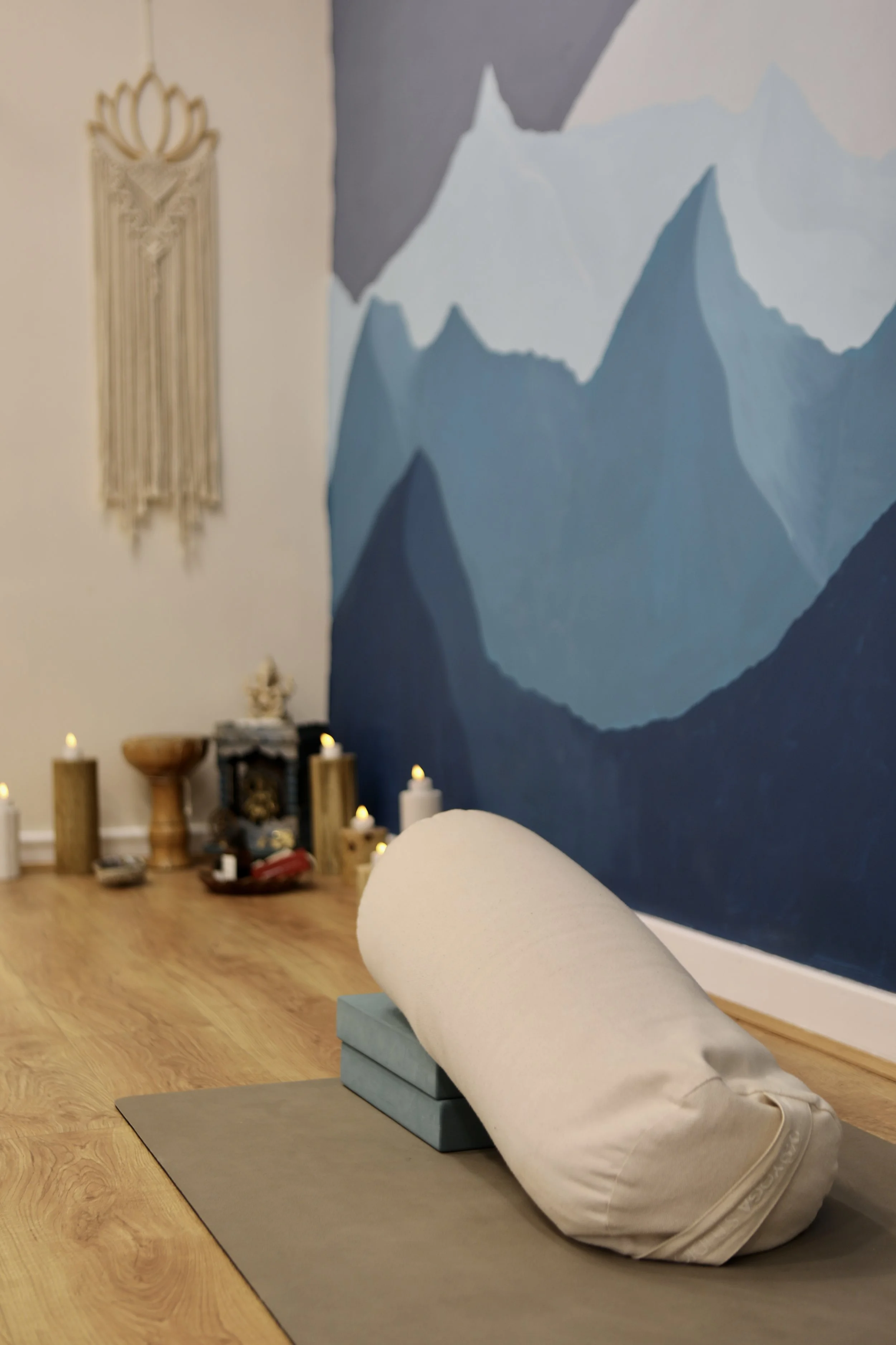 A yoga studio with a grey mat and a bolster pillow. In the background, a blue mountain mural and lit candles create a calming atmosphere.