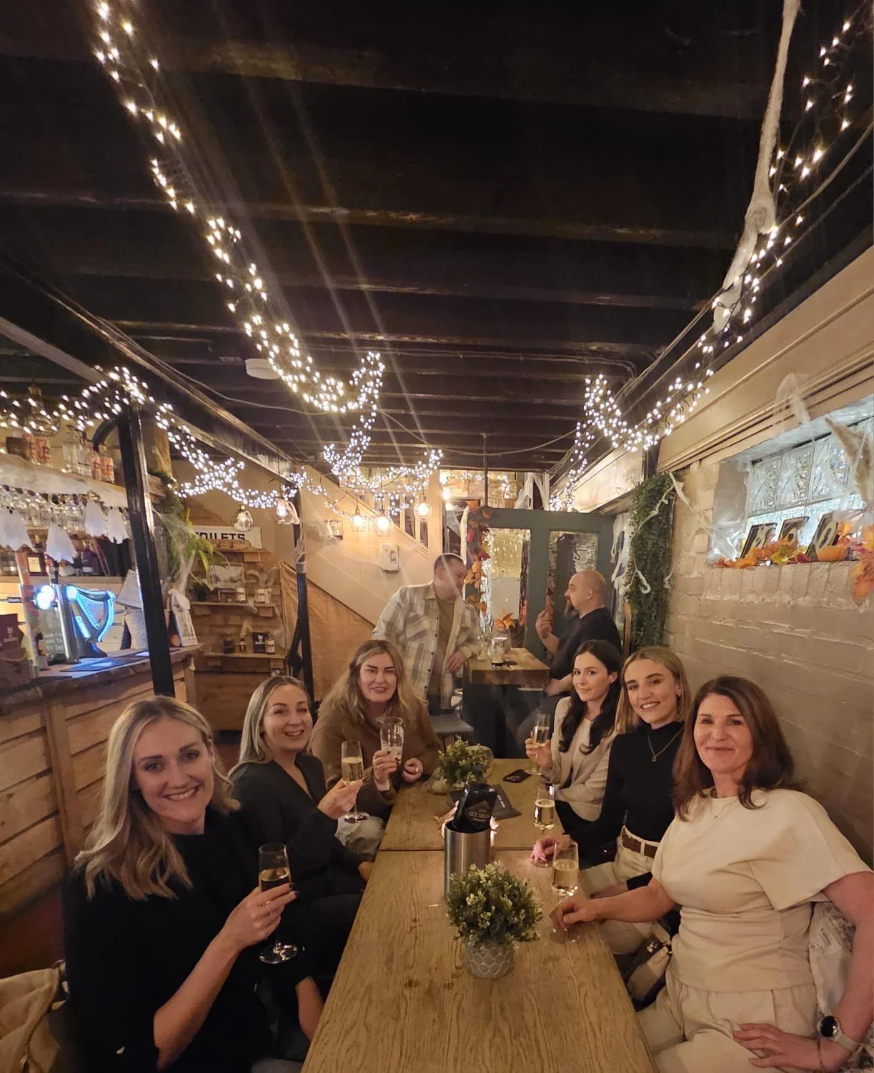 A group of women sitting around a wooden table with drinks, smiling at a cozy indoor celebration decorated with string lights and autumn-themed decorations.
