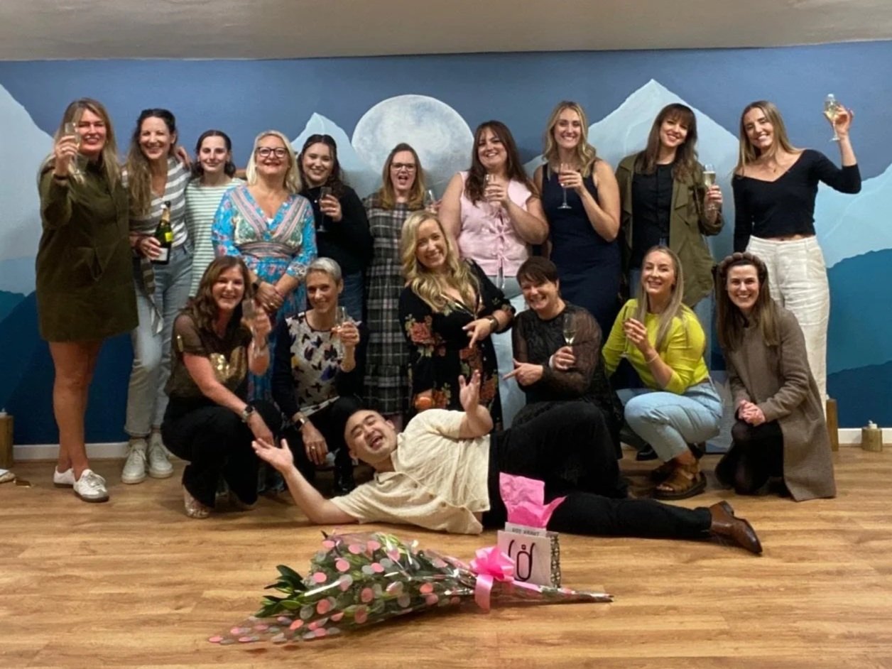 Group of women celebrating with drinks in a room with mountain mural, with a person lying in front and a bouquet of flowers on the floor.