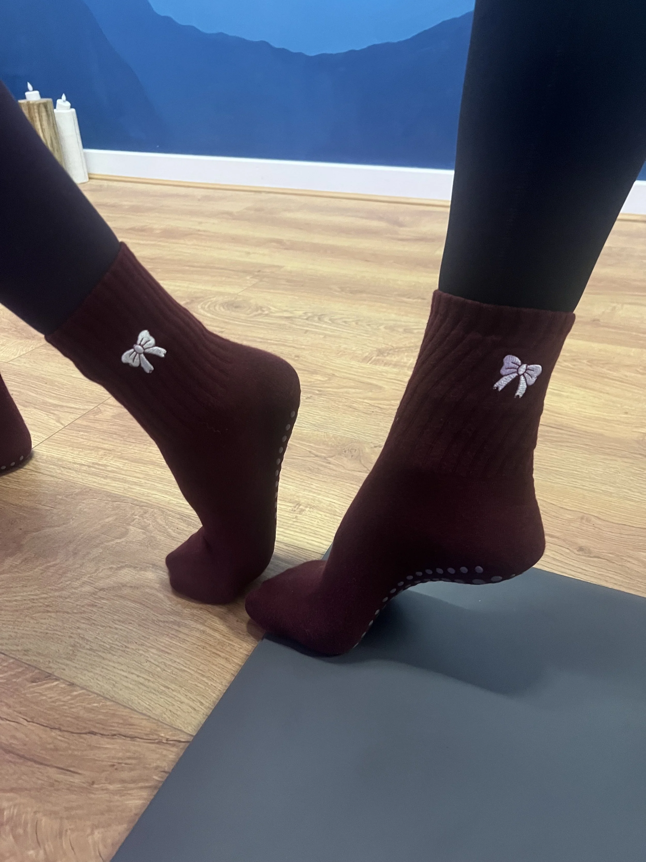 Close-up of a person's feet wearing maroon socks with white bow embroidery, standing on a gray yoga mat on a wooden floor in a room with blue mountain-themed wall art.