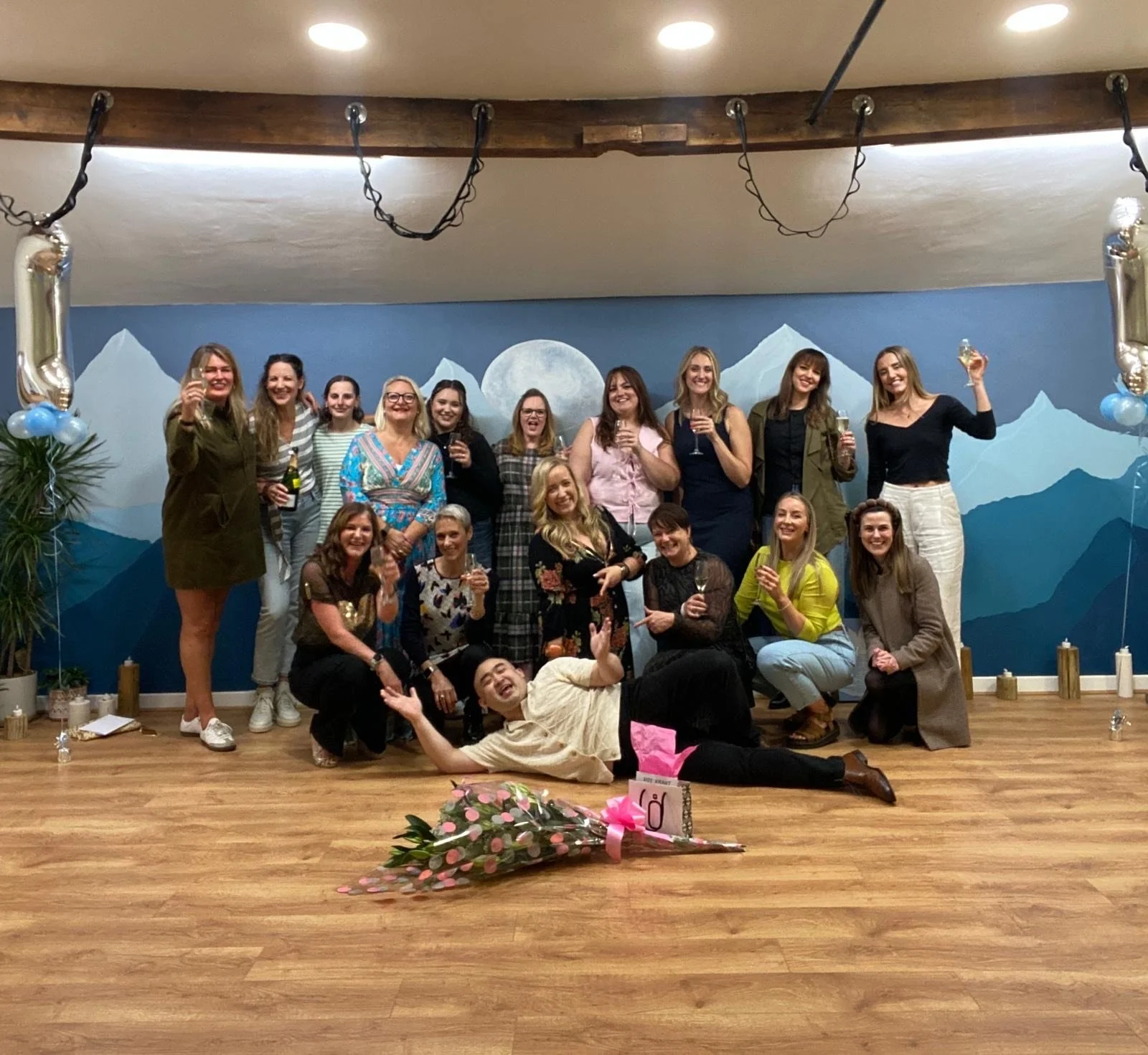 A group of women celebrating indoors with drinks, smiling, and posing for a photo in front of a mountain landscape mural. There are balloons and flowers, and a man lying on the floor in front holding a pink bouquet.