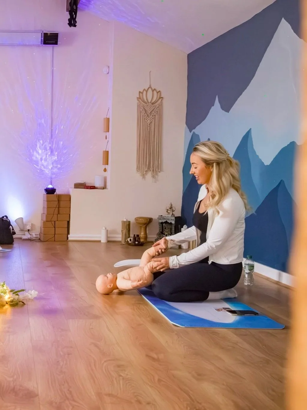 A woman practicing infant massage on a doll in a room with a blue mountain mural, wooden floor, and decorative wall hanging.