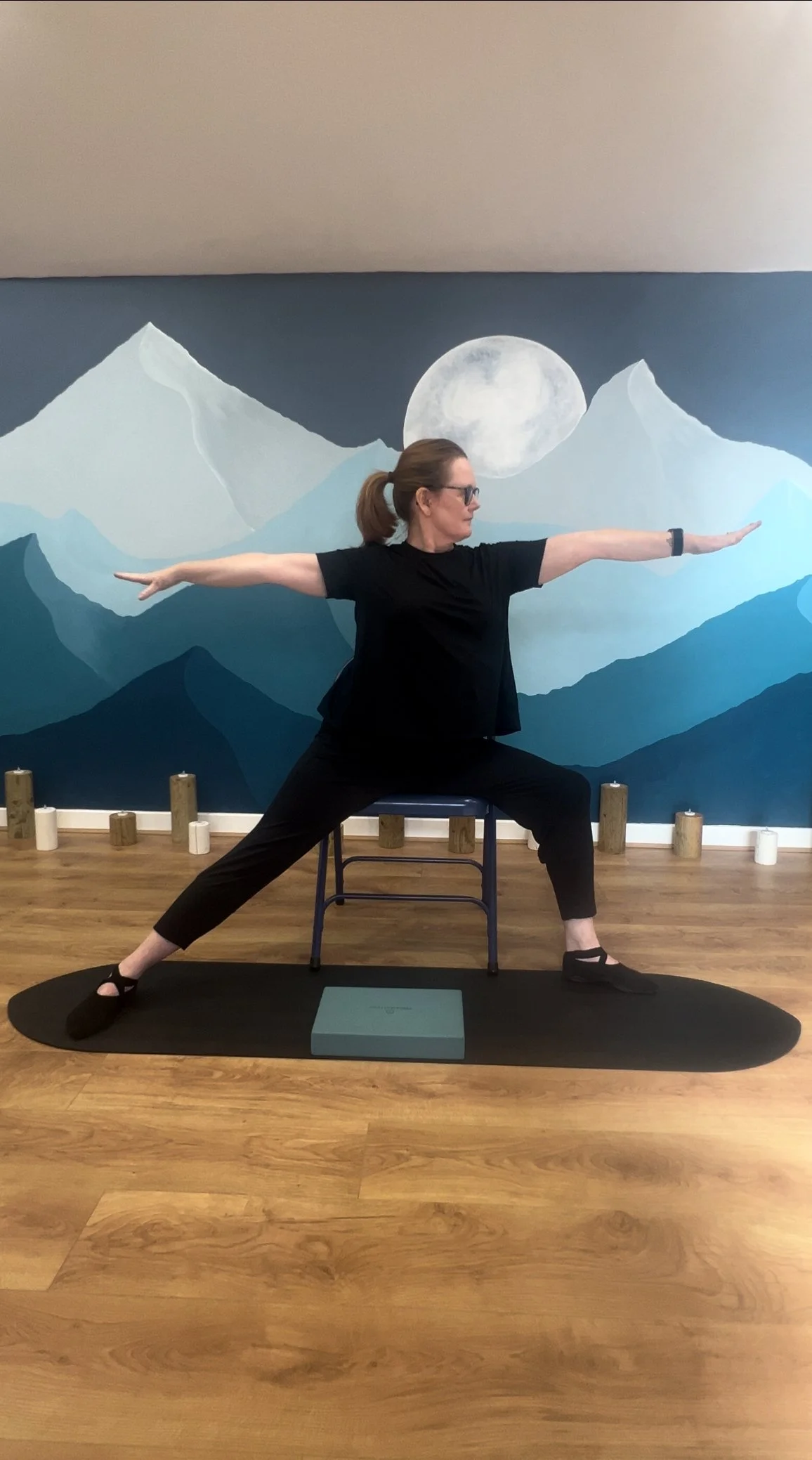 Woman practicing yoga in a lunge pose with arms extended, seated on a chair on a yoga mat in front of a mountain mural.