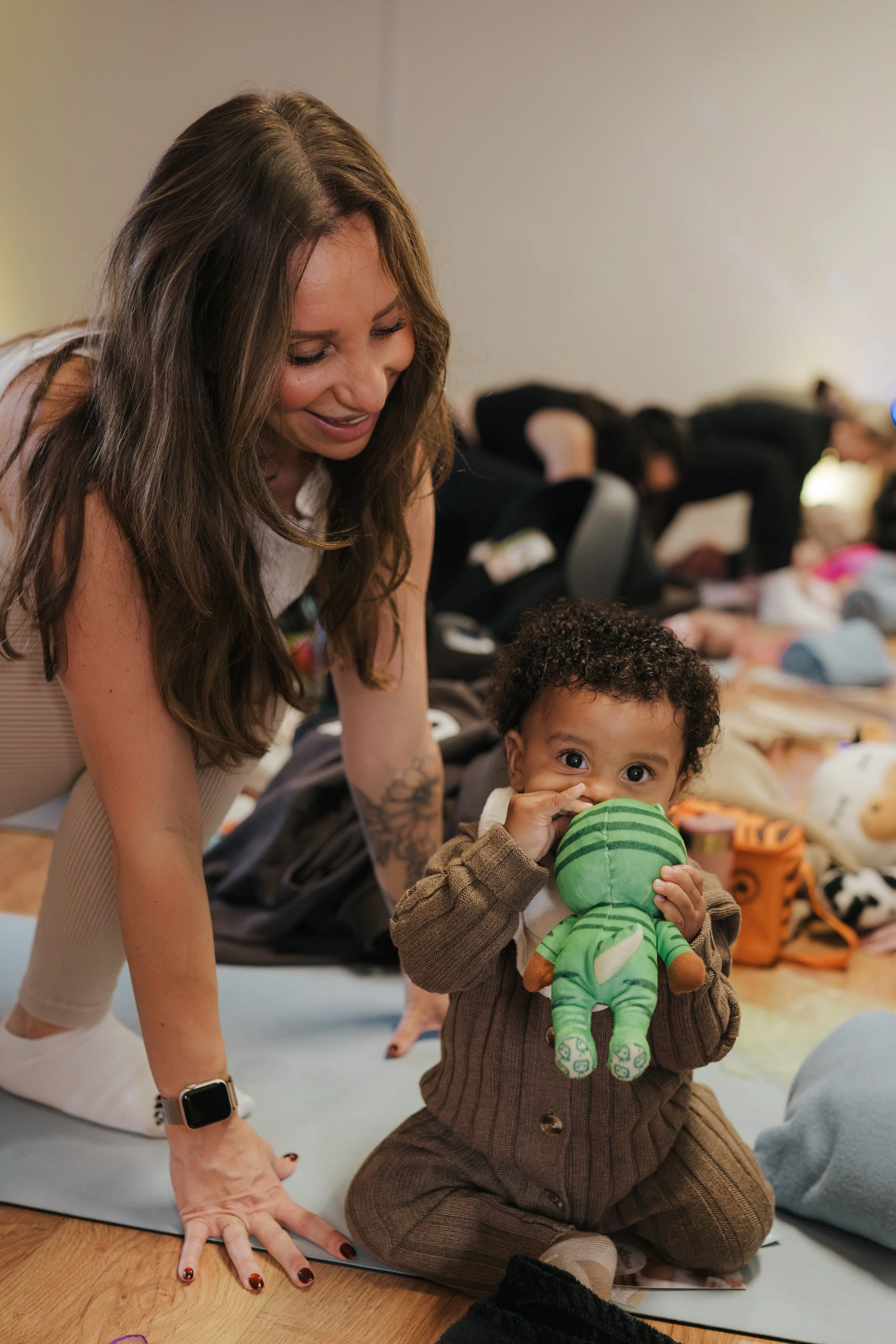A woman and a young child on a yoga mat, with the woman smiling and the child holding a green stuffed toy, surrounded by other people and toys in the background.
