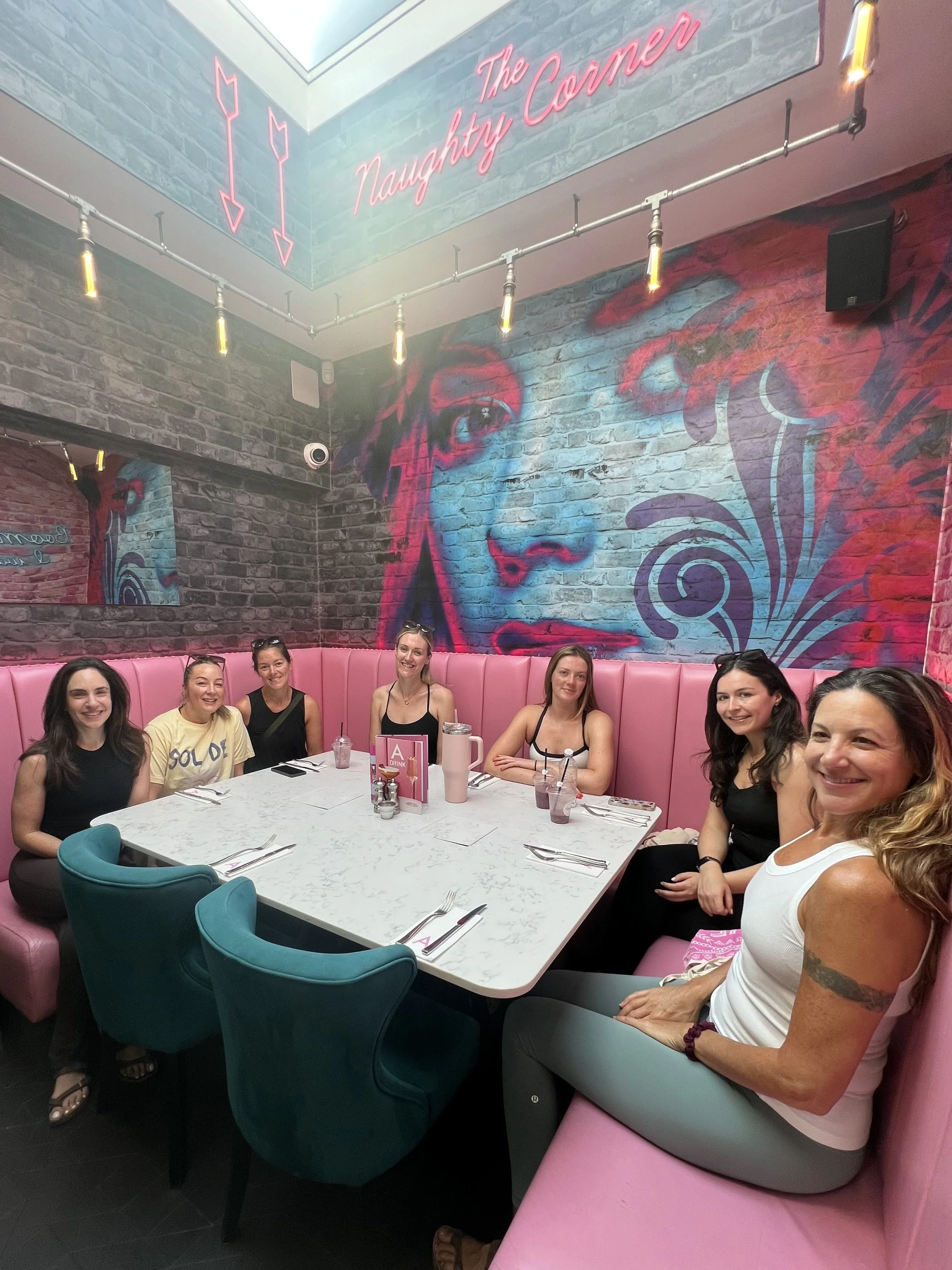 A group of seven women sitting around a white table in a neon-decorated restaurant called "The Naughty Corner." The restaurant has a mural of a woman's face on the brick wall and pink seating.