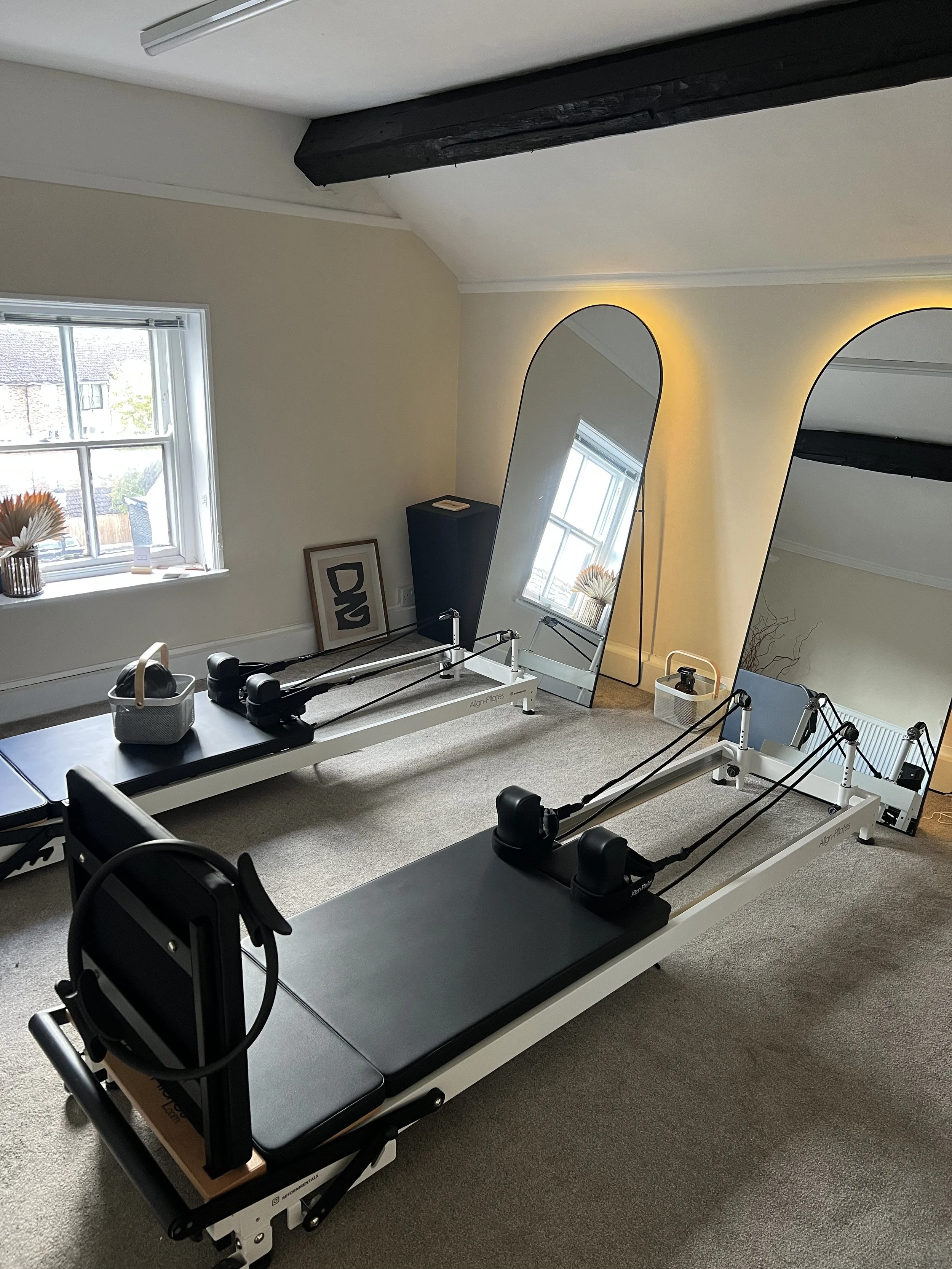 Two Pilates reformer machines in a room with beige carpet, a window with a vase of flowers on the sill, a mirror leaning against the wall, and a framed artwork.