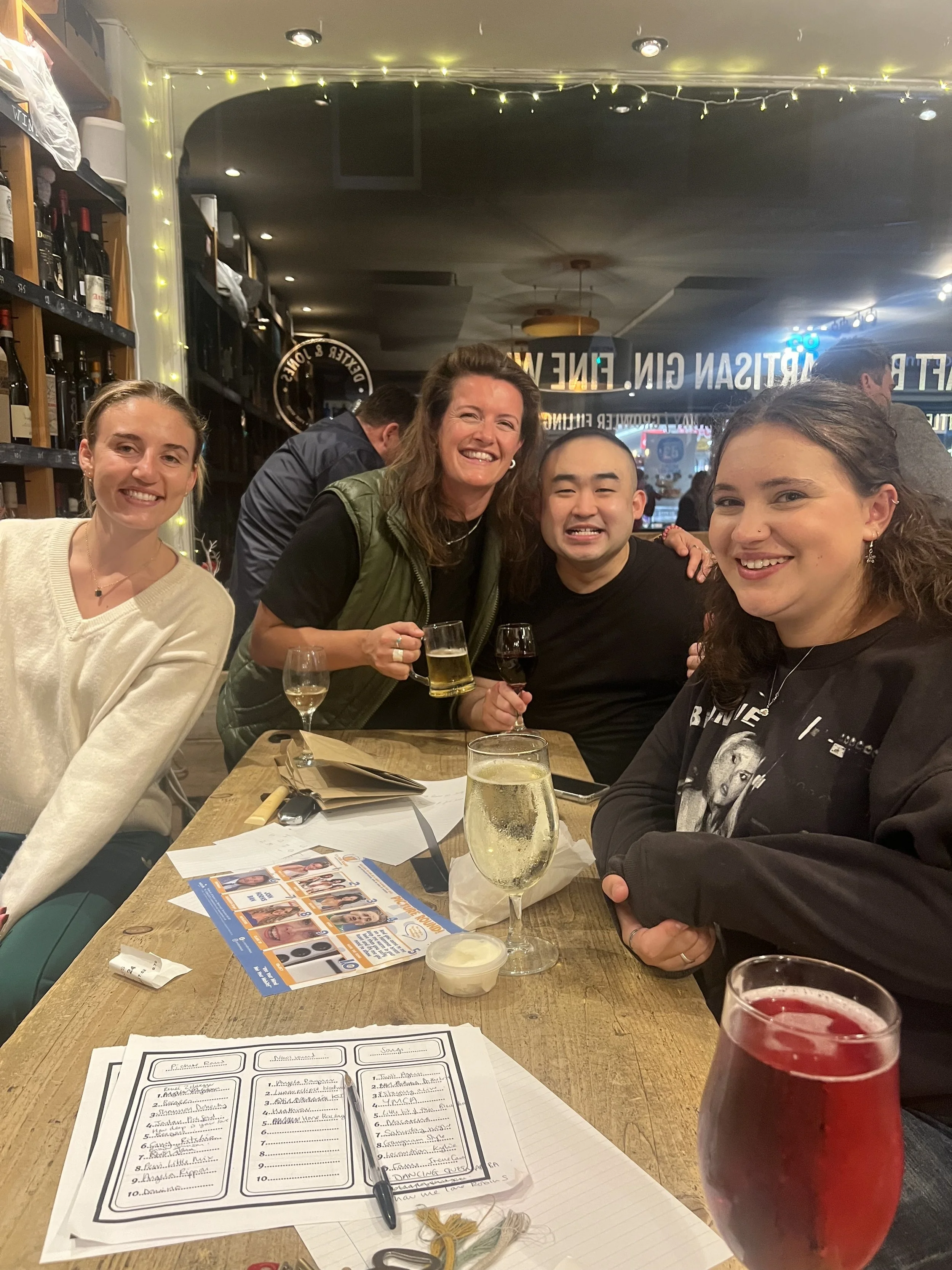 Group of four people smiling and posing for a photo at a restaurant table with drinks, menus, and napkins. The background shows a bar and a window with city views.