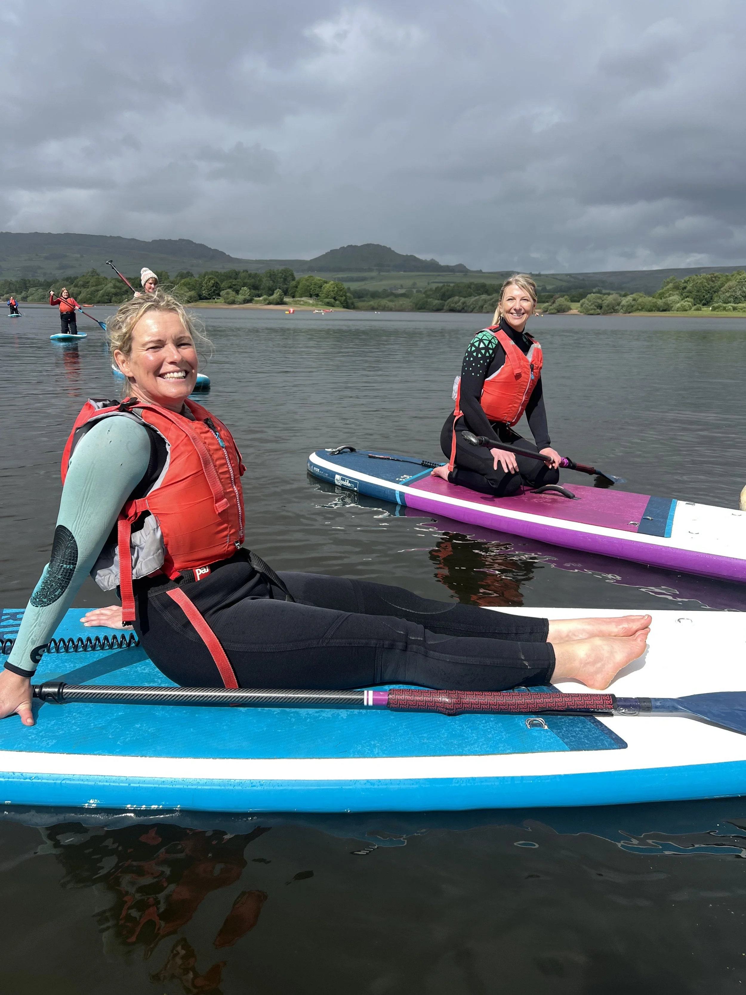 Two women in life jackets on paddleboards enjoying a day on a calm lake with overcast sky and green hills in the background.