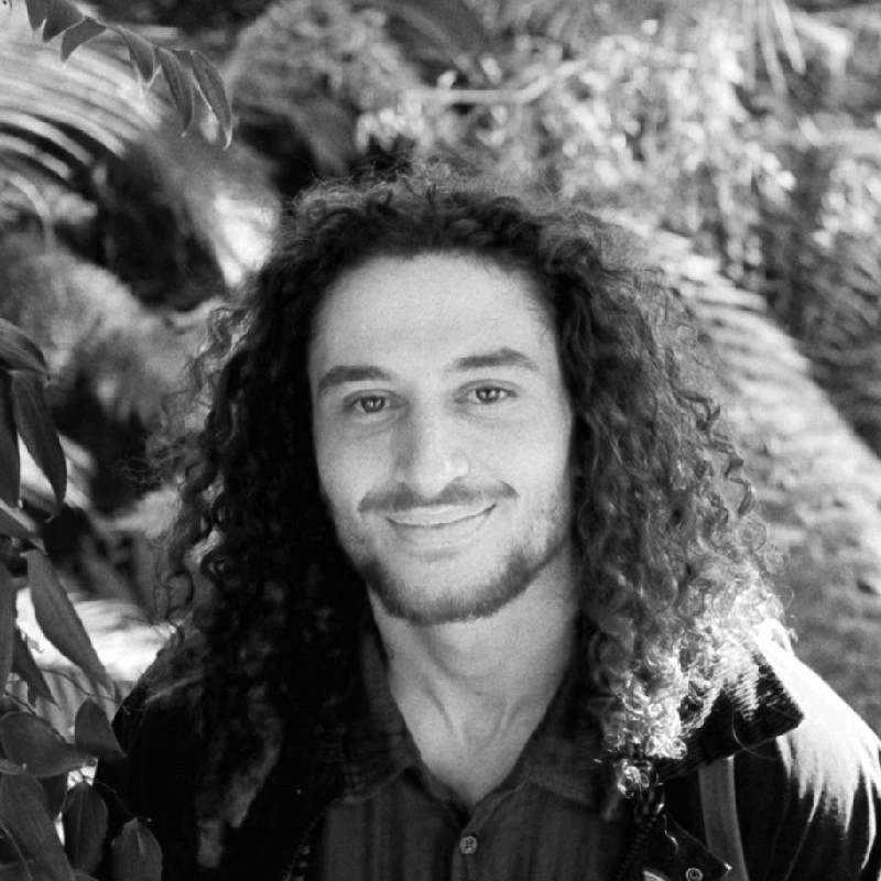 A young man with long, curly hair smiling outdoors among foliage.