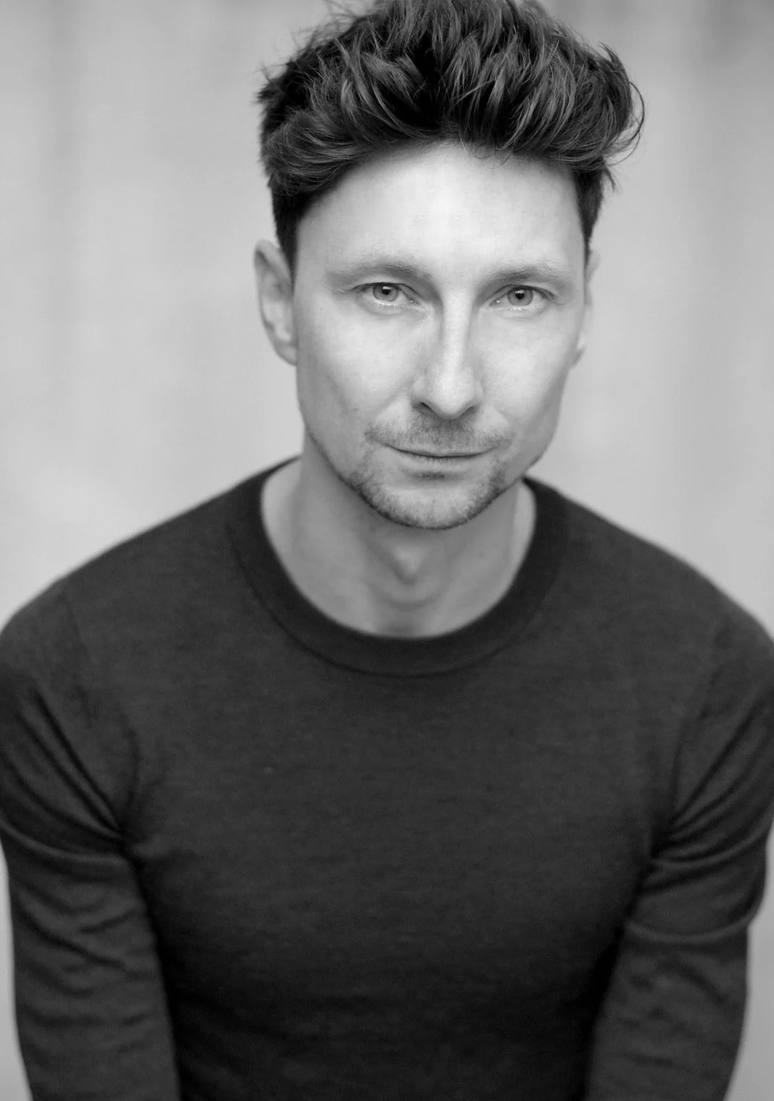 A black and white portrait photograph of a man with short hair and a neutral expression, wearing a dark jumper.