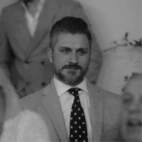 A man with a dark beard and mustache wearing a light-colored suit and a polka dot tie, sitting indoors.