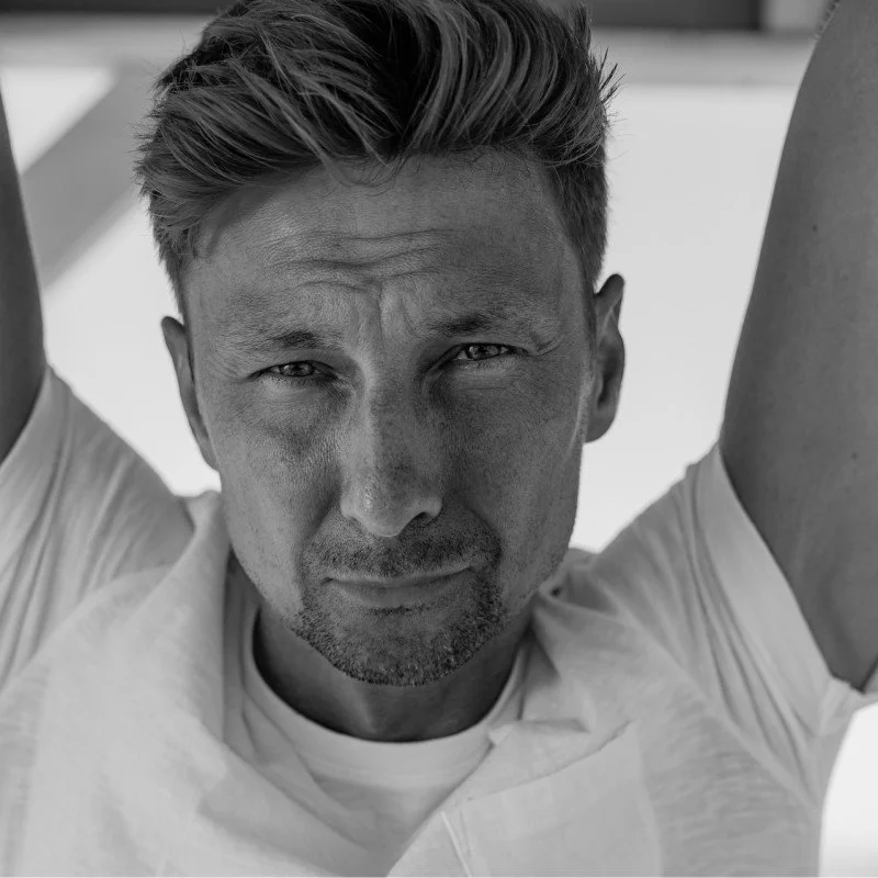 A black and white close-up of a man with short hair and a serious expression, wearing a white t-shirt.