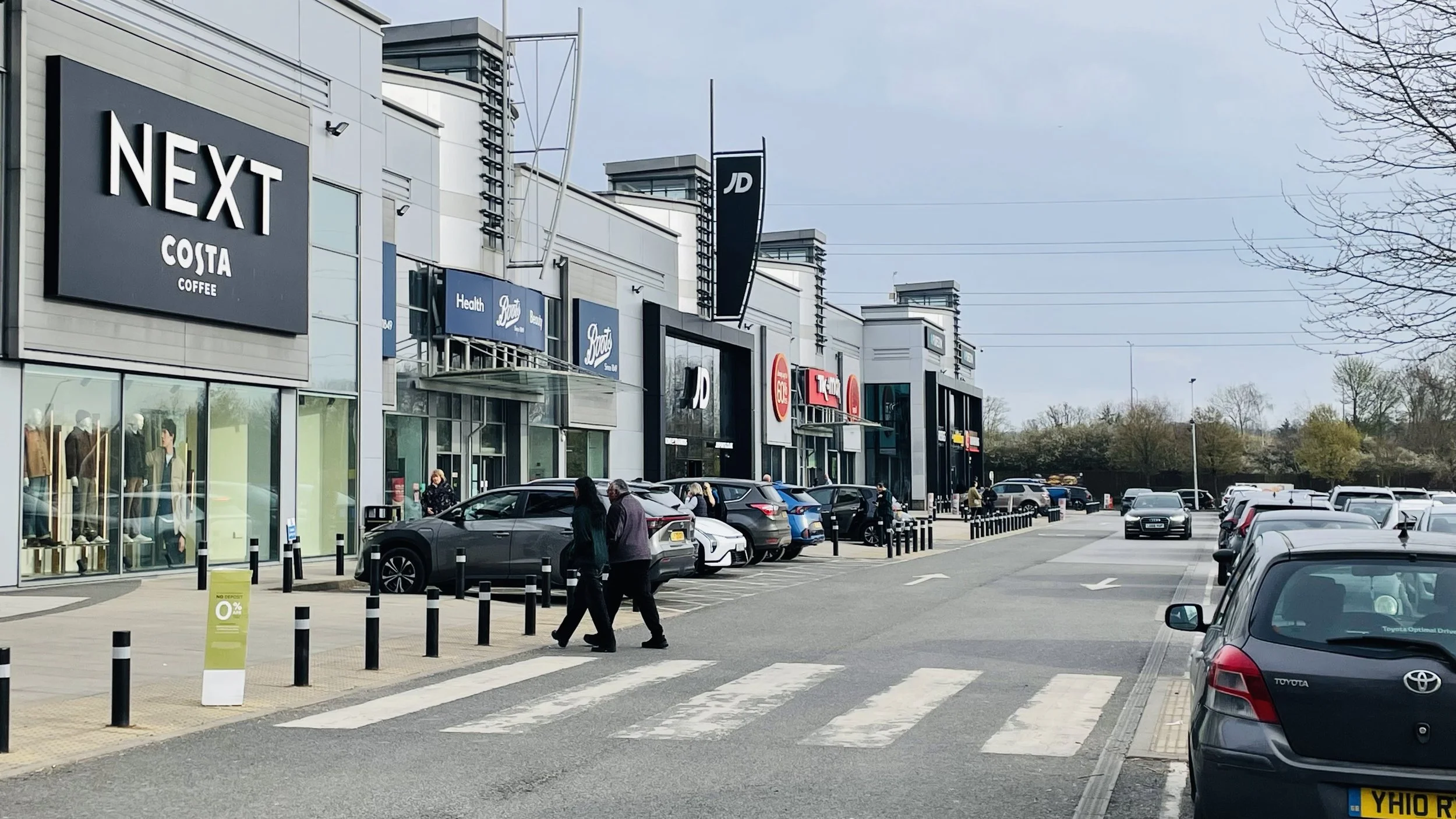 An image of a retail park featuring a line of shops including Next, Costa, Boots, JD Sports and TK Maxx, next to a car park.