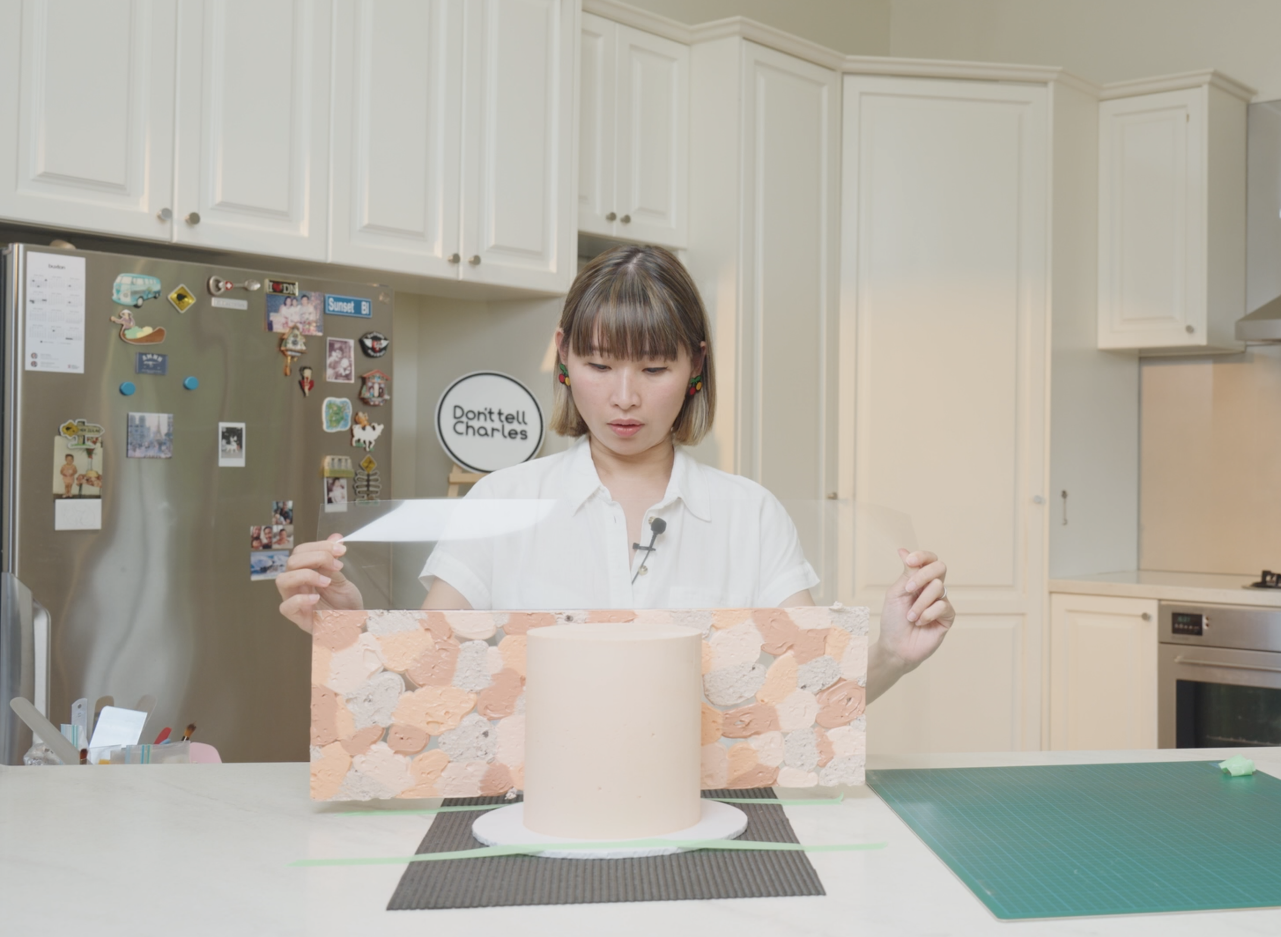 A woman in a white shirt is working on a cake with a patterned panel on a kitchen countertop. The kitchen has white cabinets and a stainless steel fridge decorated with various magnets. There is a sign that says 'Don't tell Charles' on an easel in the background.