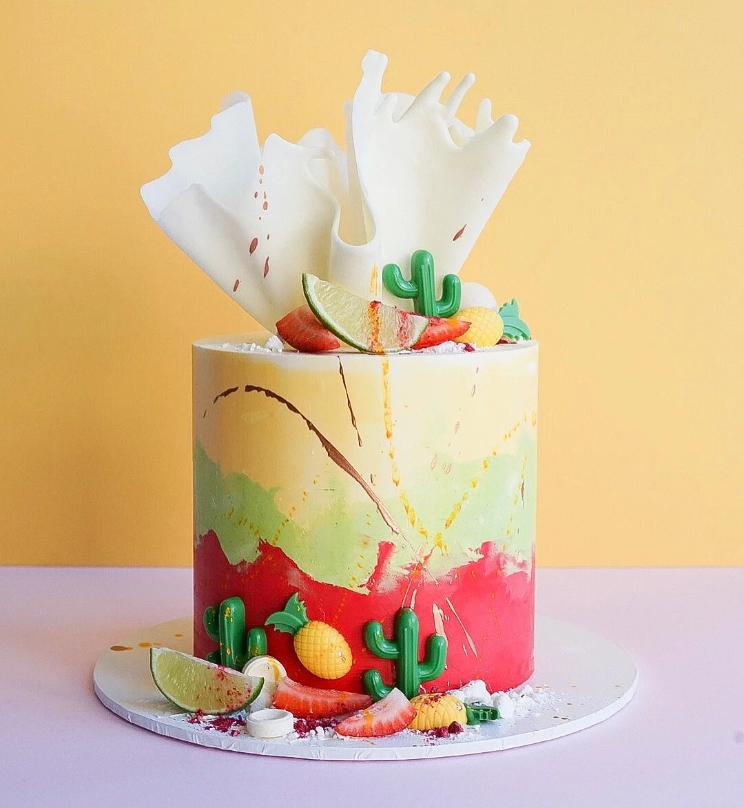 Colorful desert-themed cake with cactus, lime wedge, strawberries, pineapple, and decorative white chocolate pieces, set against yellow background.