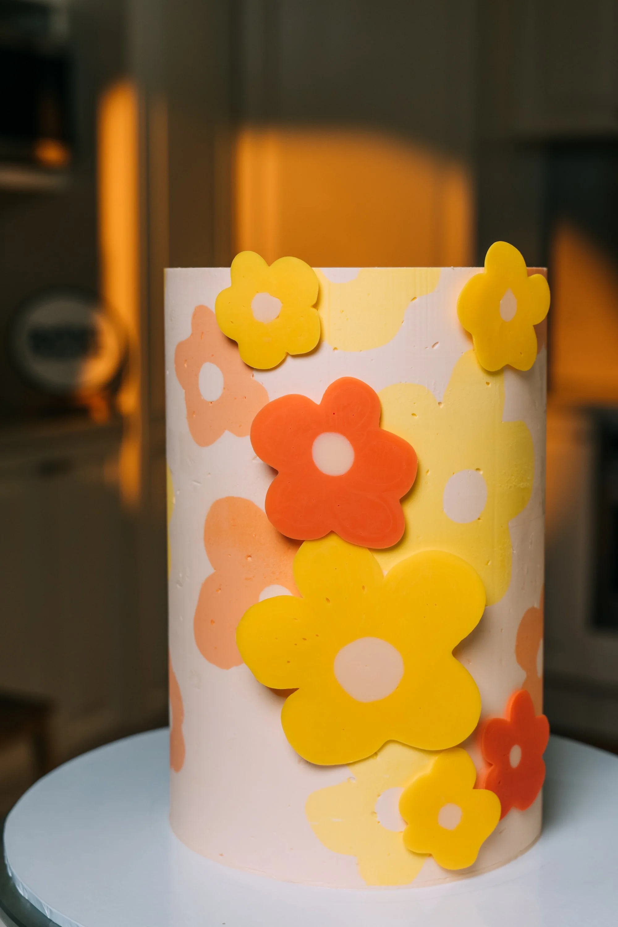 A white cake decorated with colorful flower-shaped fondant flowers in yellow, orange, and peach colors.