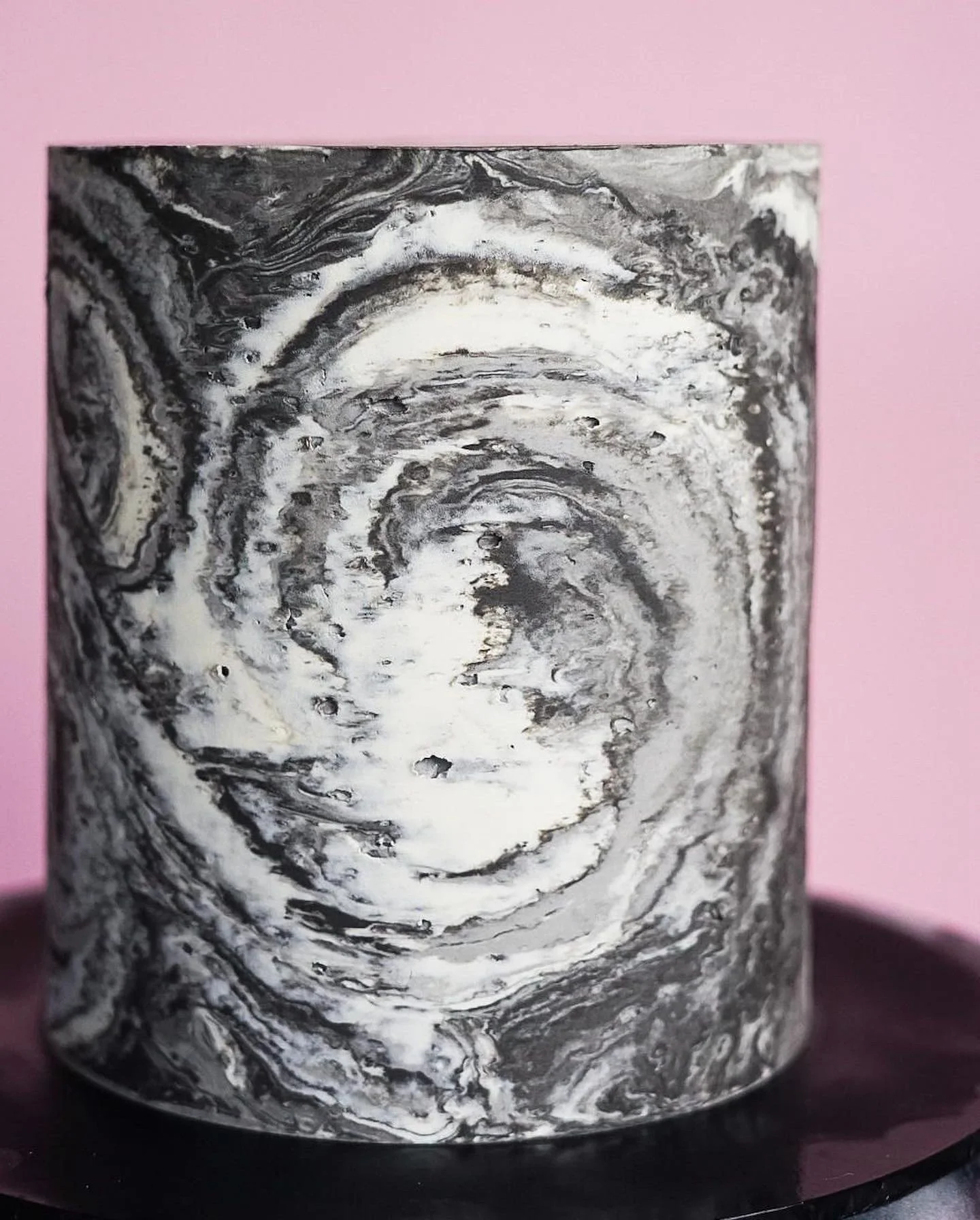 Marble-patterned cylindrical object with black, white, and gray swirls sitting on a black surface against a pink background.