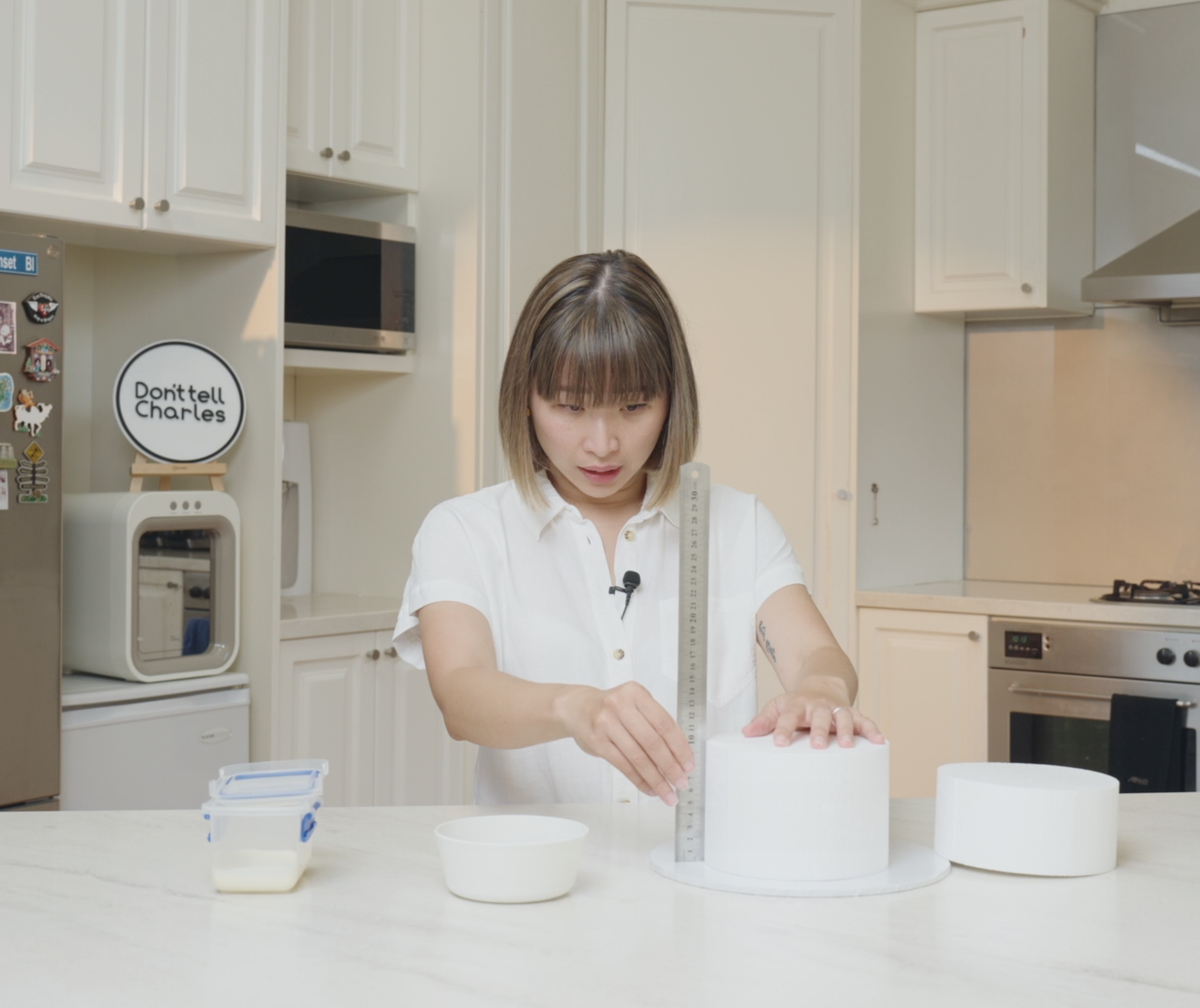 A woman in a white shirt is measuring a block with a ruler in a bright kitchen with white cabinets. Various kitchen items, including a small container of milk, are on the counter.
