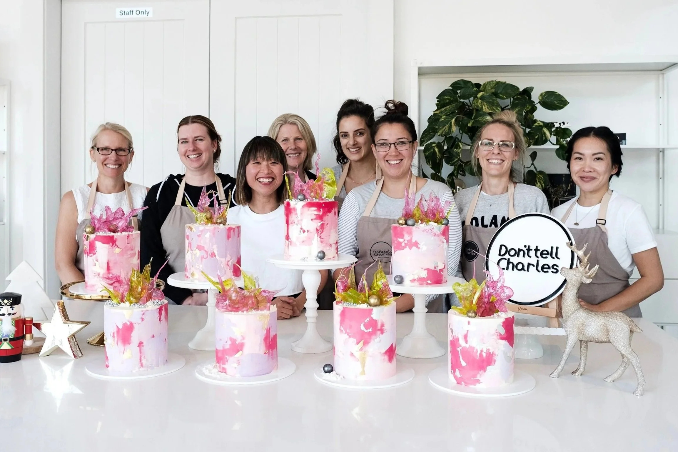 Group of women in aprons smiling behind decorated cakes at a celebration.