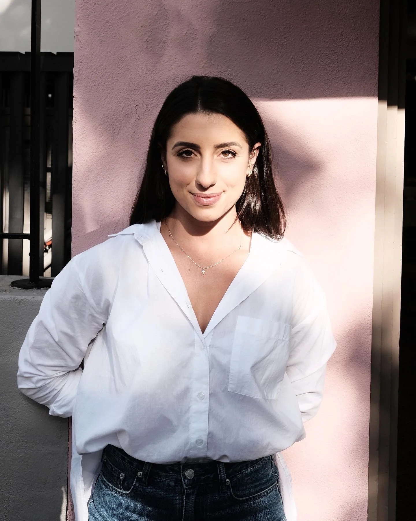 A young woman with dark hair and light skin, wearing a white shirt and dark jeans, standing outdoors against a pink wall with sunlight and shadow.